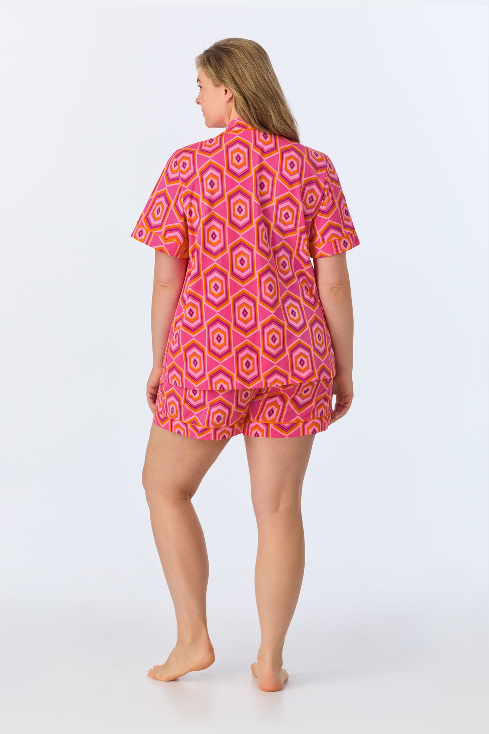 A woman stands barefoot with her back to the camera, modeling the TT SS Shorty PJ Set - MLT Hexagon Jersey against a plain white background. She wears the geometric-patterned pink and orange short-sleeve shirt with matching shorts.