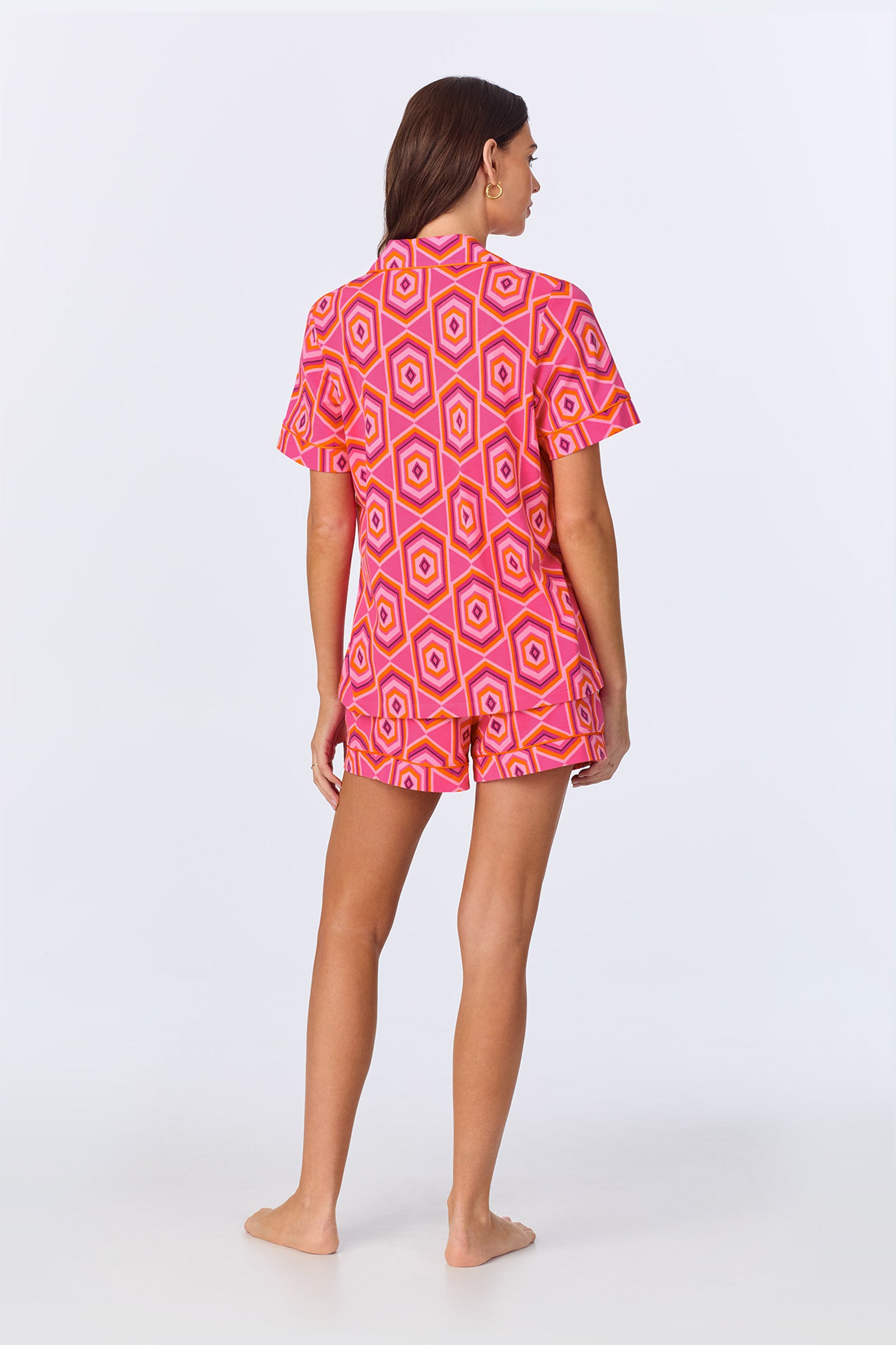 A woman stands barefoot facing away, wearing the TT SS Shorty PJ Set - MLT Hexagon Jersey, featuring a vibrant pink and yellow geometric pattern, against a plain white background.