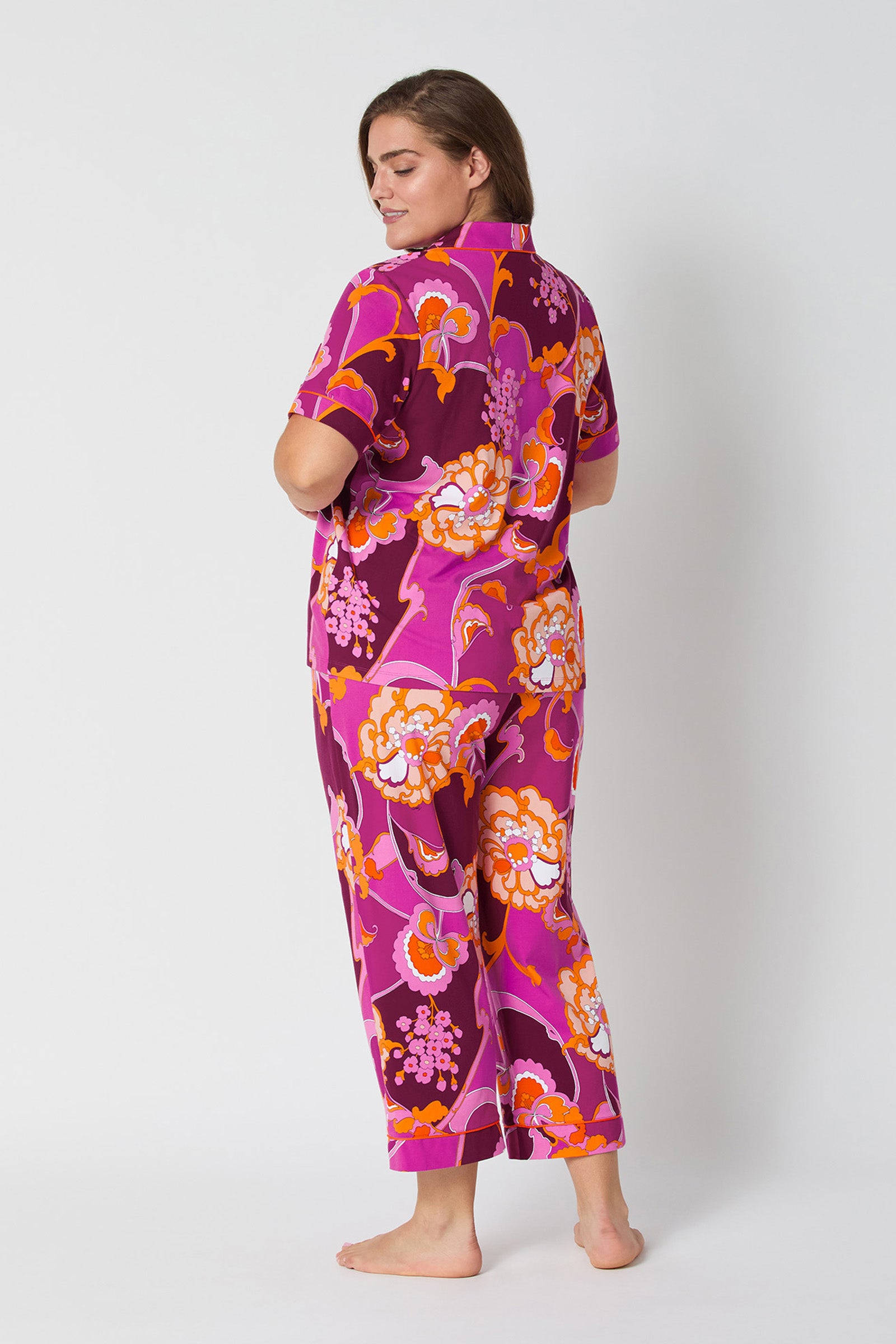 A woman stands barefoot, facing away and smiling gently, wearing the Apache Plume S/S Cropped Pj—a vibrant pink pajama set with bold orange, red, and white floral patterns on both the cropped top and pants.