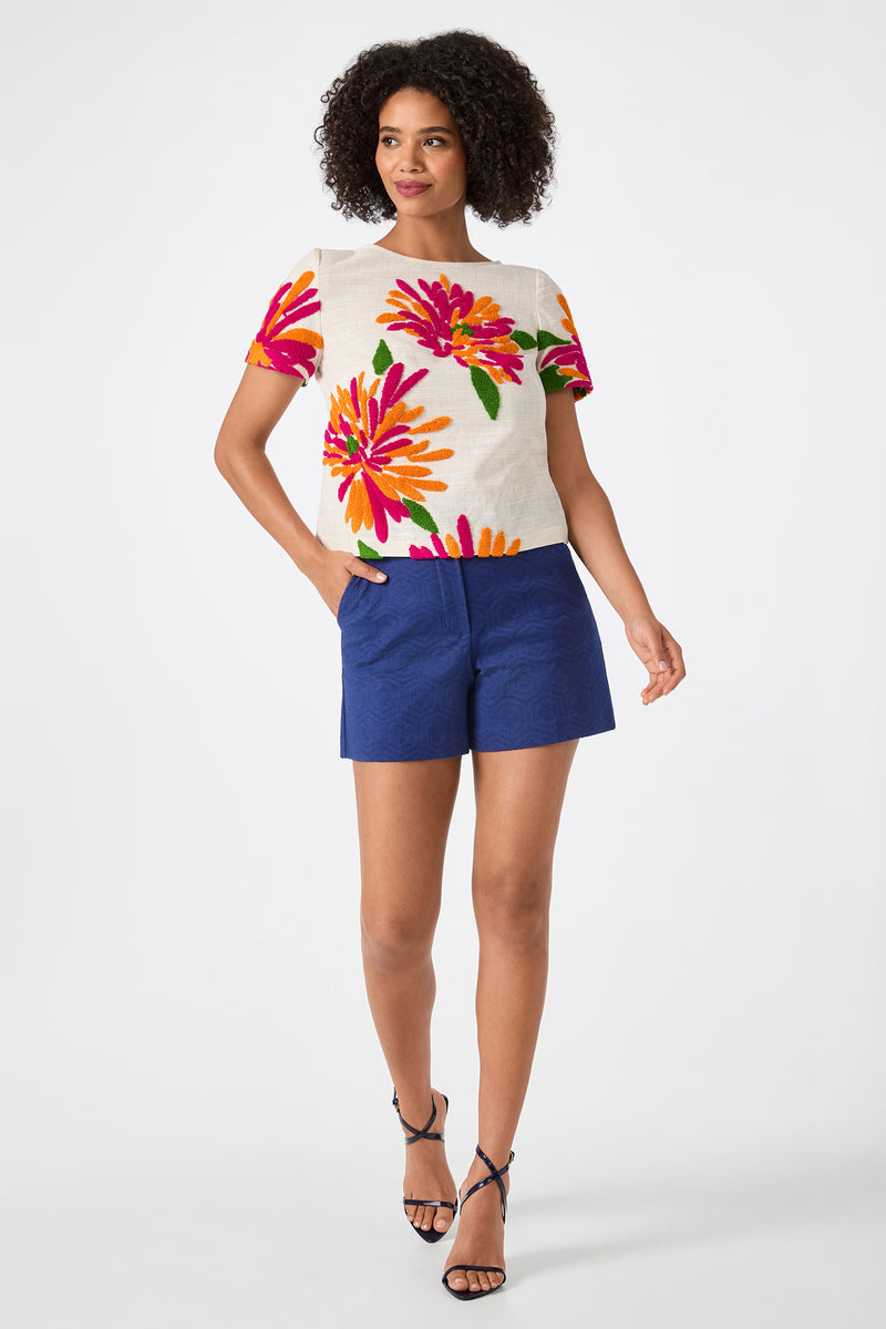 A woman with curly hair smiles against a white background, wearing the Valera Shorts - Jin Jacquard in blue, a white floral short-sleeve top, and black strappy heels.
