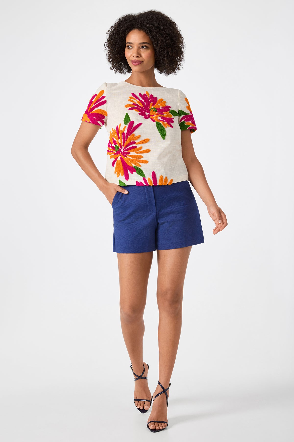 A woman with curly hair smiles against a white background, wearing the Valera Shorts - Jin Jacquard in blue, a white floral short-sleeve top, and black strappy heels.