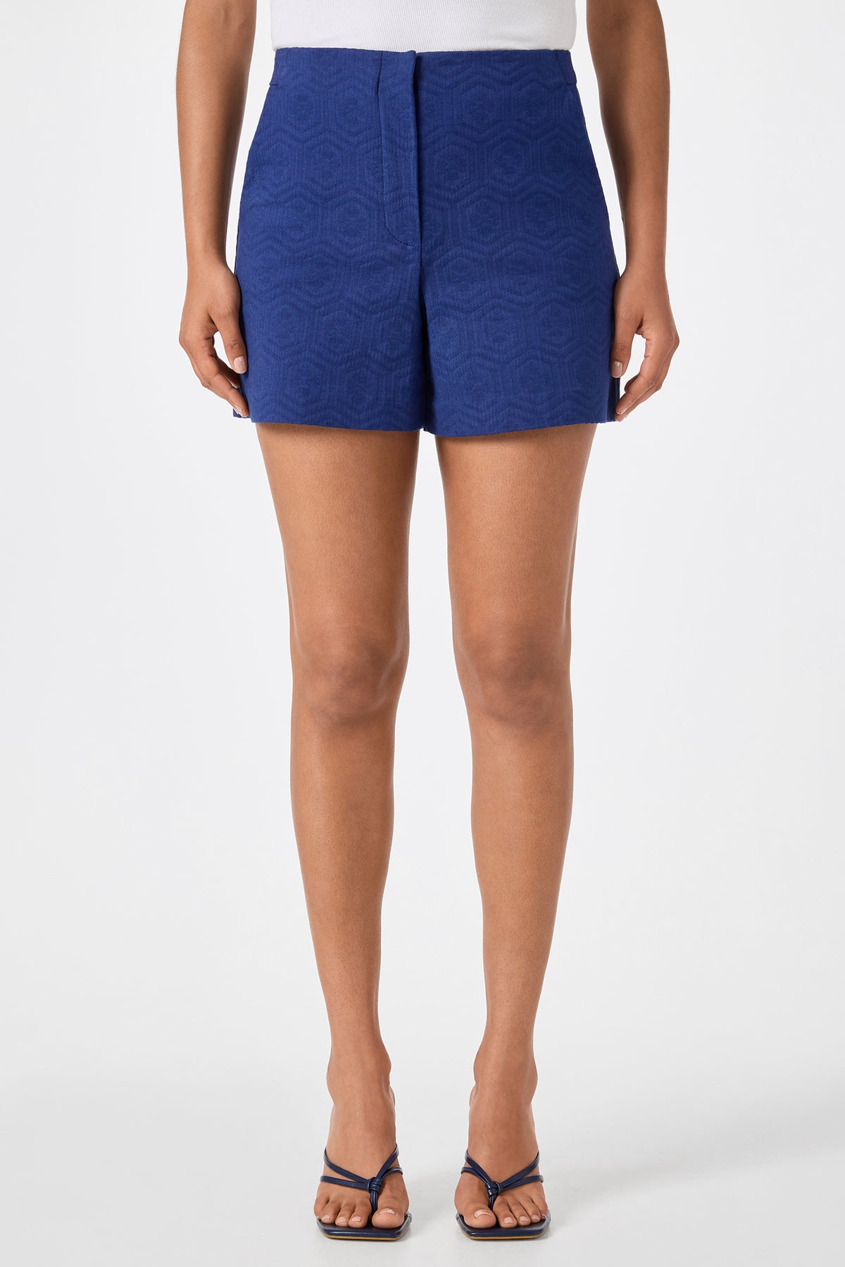 A person wearing a white top, Valera Shorts - Jin Jacquard in Noh Nightfall, and strappy heeled sandals stands against a plain white background.