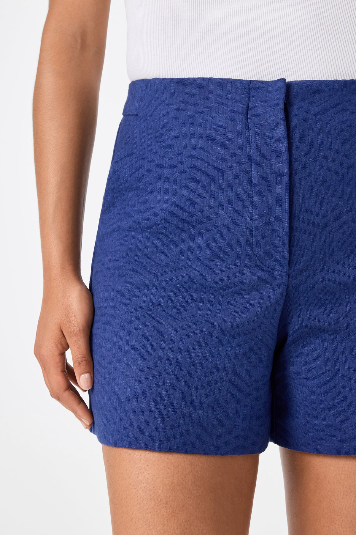 A person wearing Valera Shorts - Jin Jacquard, which are textured, high-waisted royal blue shorts, paired with a white ribbed top. Only their midsection, right arm, and upper thighs are shown.