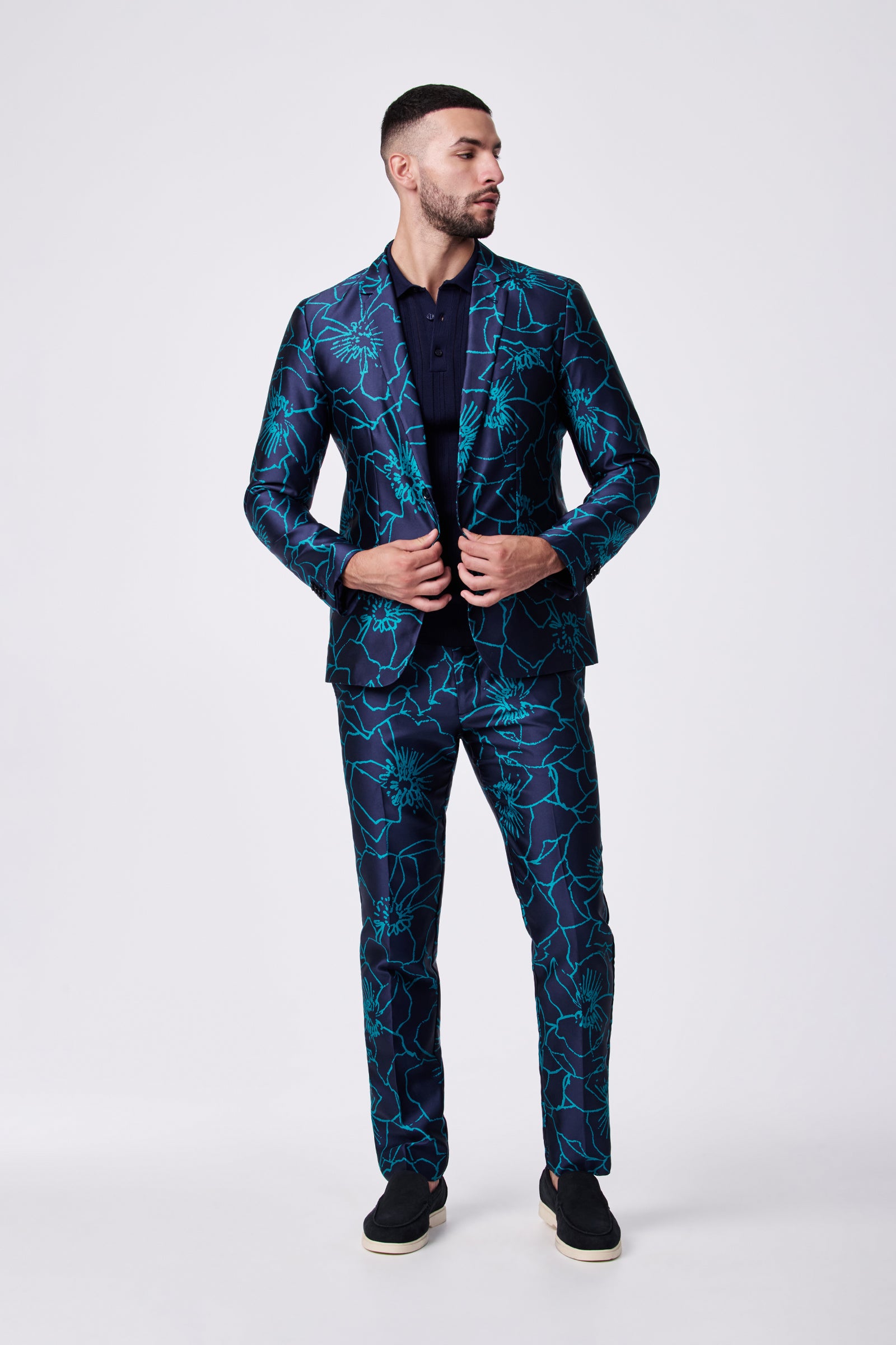 A man looks to the side and adjusts his suit jacket while wearing the Clyde Slim Trouser—a dark blue trouser with a bold, bright blue floral pattern—paired with a navy shirt and black slip-on shoes against a plain background.