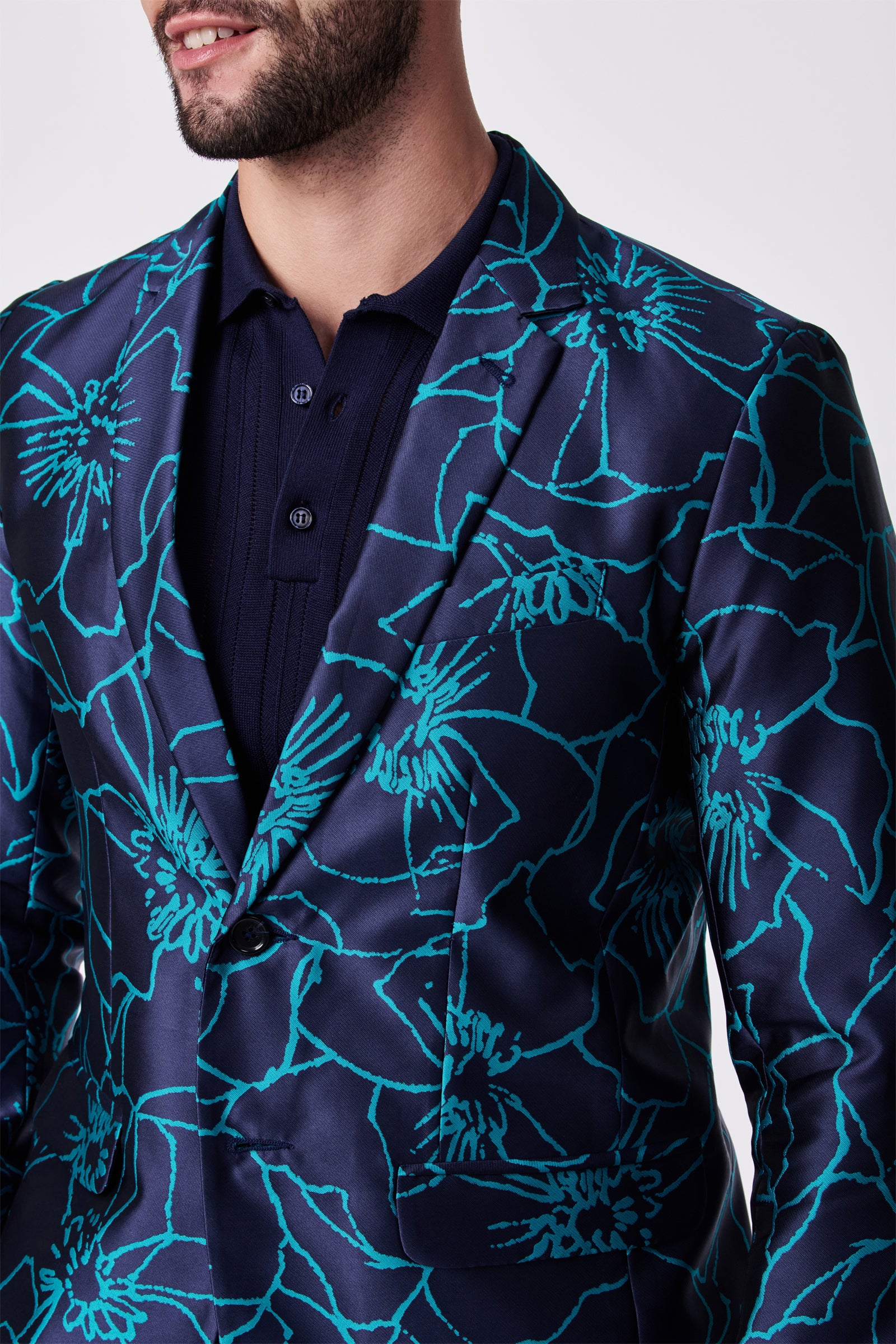 A man wearing the Thurston Blazer, which features a bold turquoise floral pattern on dark blue, over a navy blue collared shirt. He is shown from shoulders to mid-torso with part of his face visible.