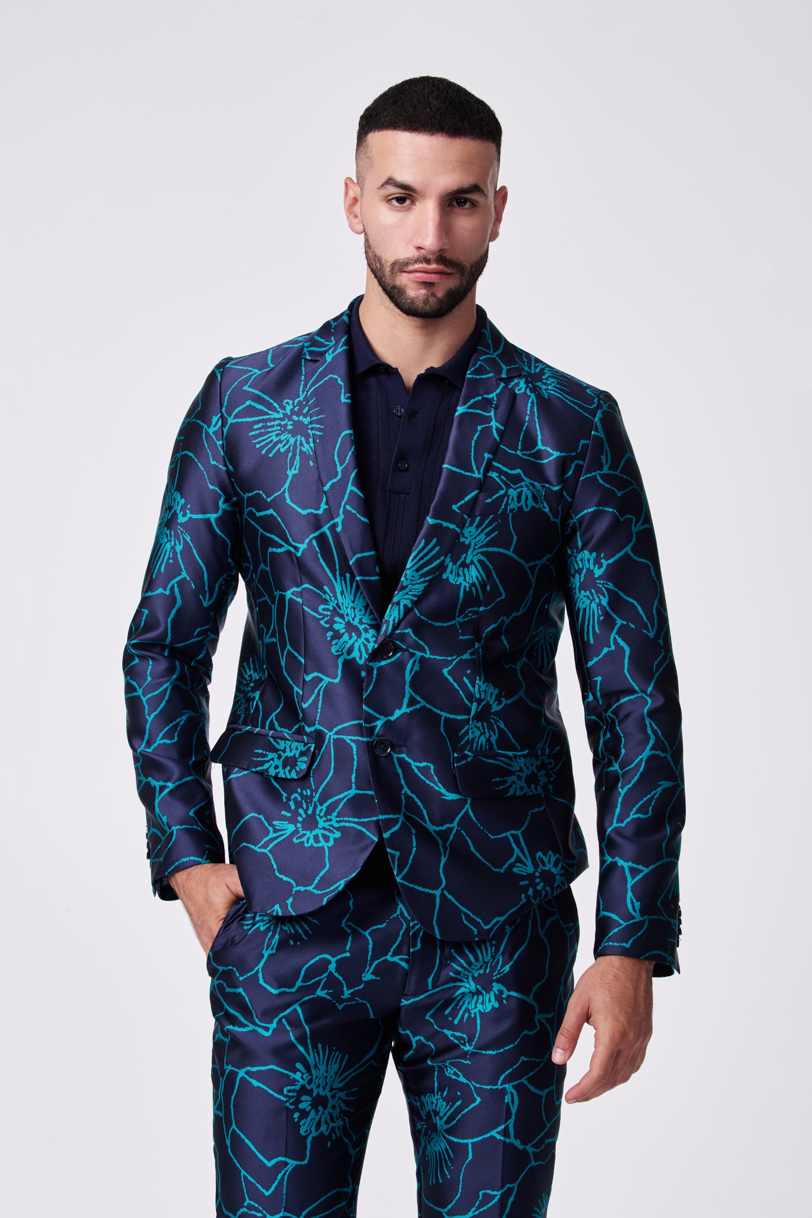 A man with short dark hair and a beard wears the Thurston Blazer, a dark blue suit with a bright blue floral pattern, standing confidently against a plain light background with one hand in his pocket.