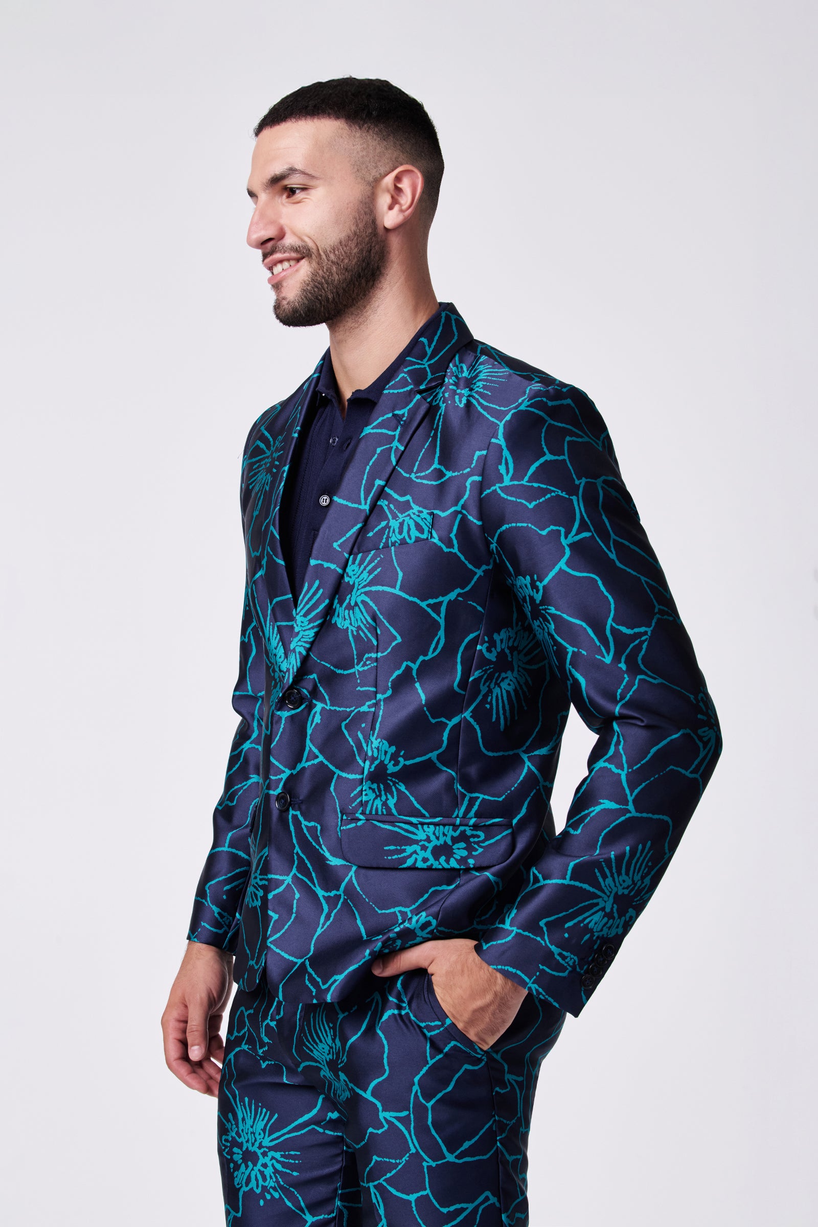 A man models the Thurston Blazer—dark blue with bold teal floral patterns—standing with one hand in his pocket, smiling slightly to the left against a plain white background.