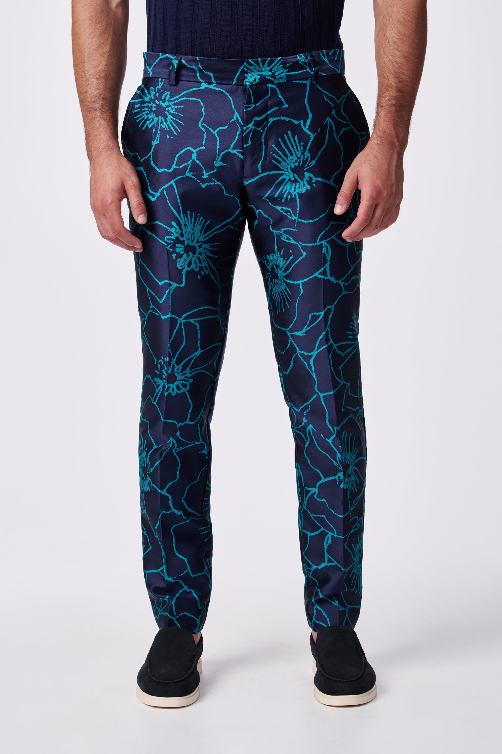 A man wears the Clyde Slim Trouser, featuring a bold teal floral pattern on navy blue, paired with black slip-on shoes and a dark shirt. The photo, cropped from shoulders down, highlights the unique design of the pants.