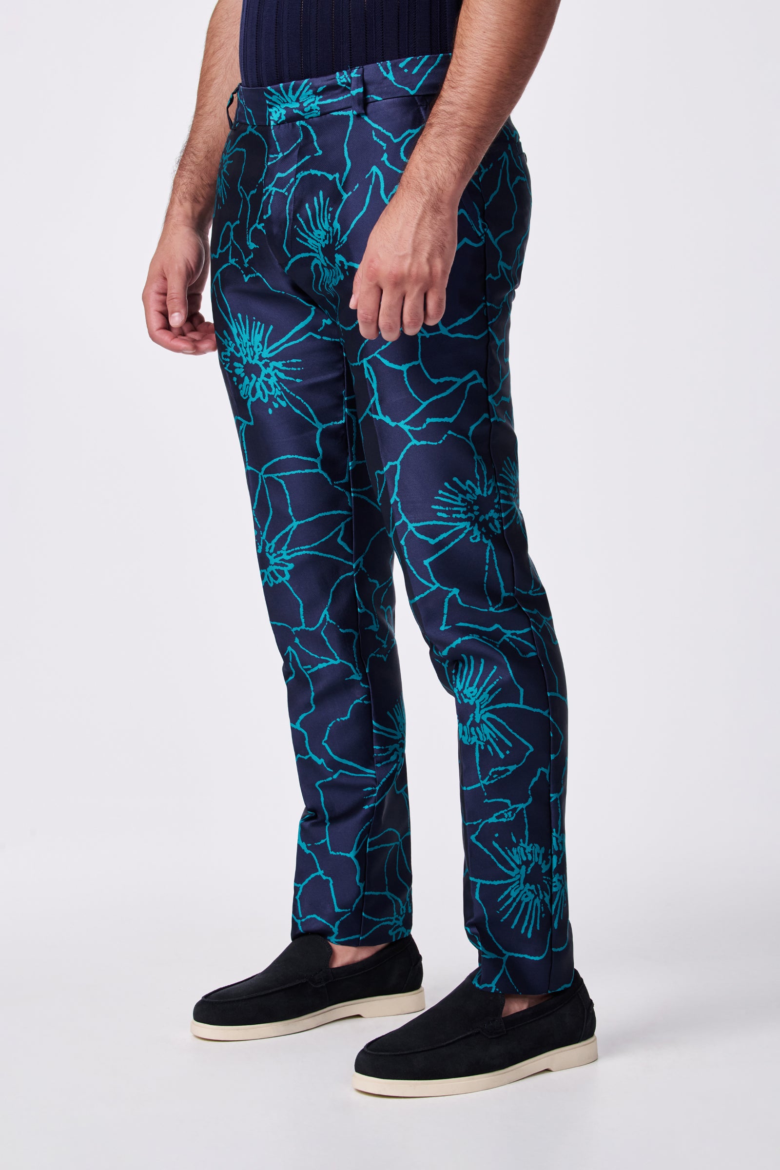 A person wearing Clyde Slim Trouser in dark blue with a bright blue floral pattern, teamed with a navy shirt and black slip-on shoes with white soles, poses against a plain white background.