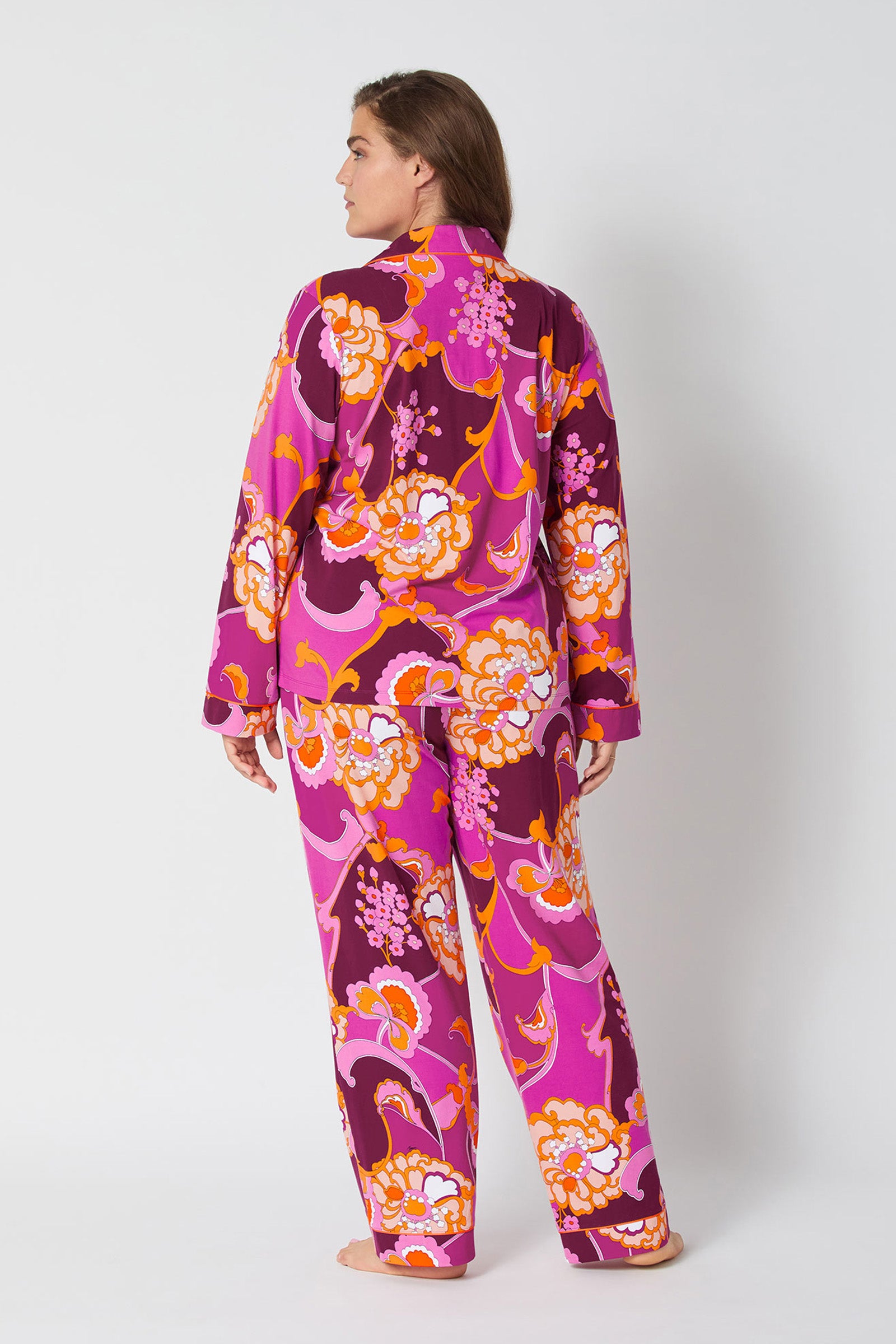 A person, seen from behind, models the Apache Plume L/S Classic Pj—a matching set with a vibrant retro floral pattern in pink, purple, orange, and white—against a plain white background.
