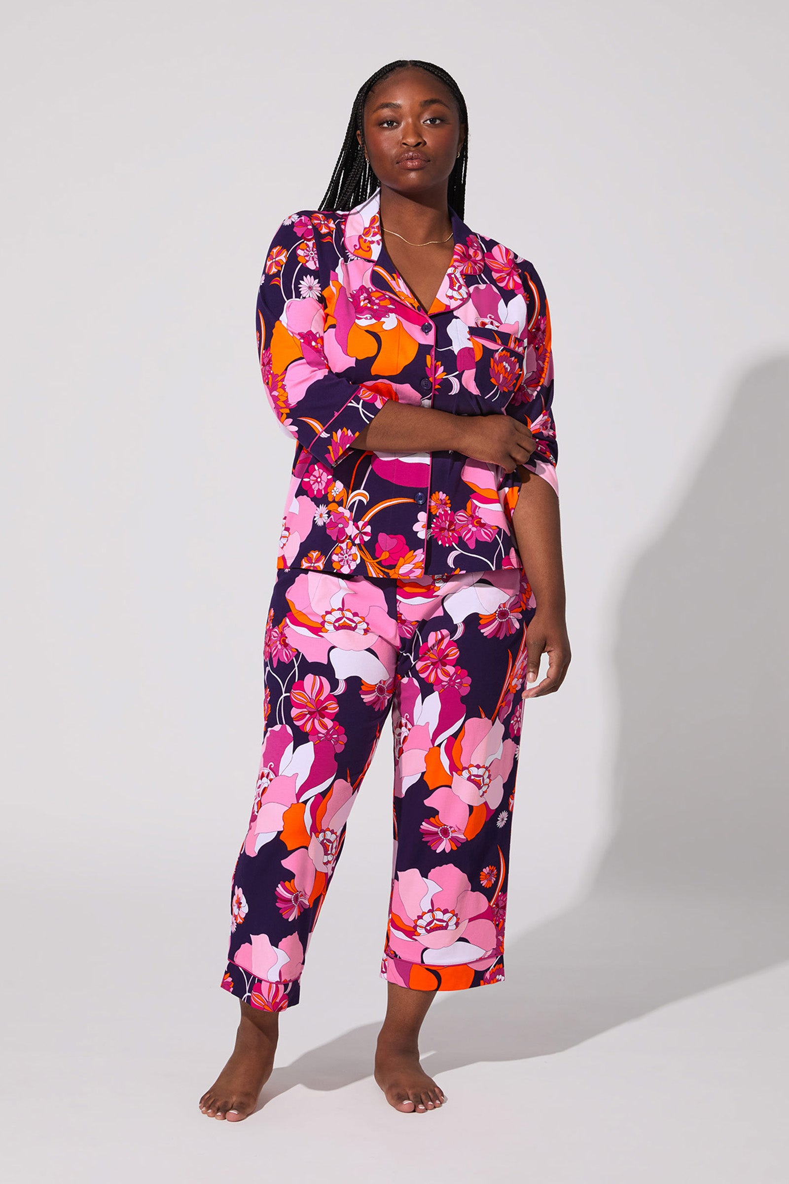 A woman with long braided hair stands barefoot against a plain background, wearing the Floral Medley Crop Pj Set featuring a bold floral pattern in pink, orange, red, and purple. She looks calm and comfortable.