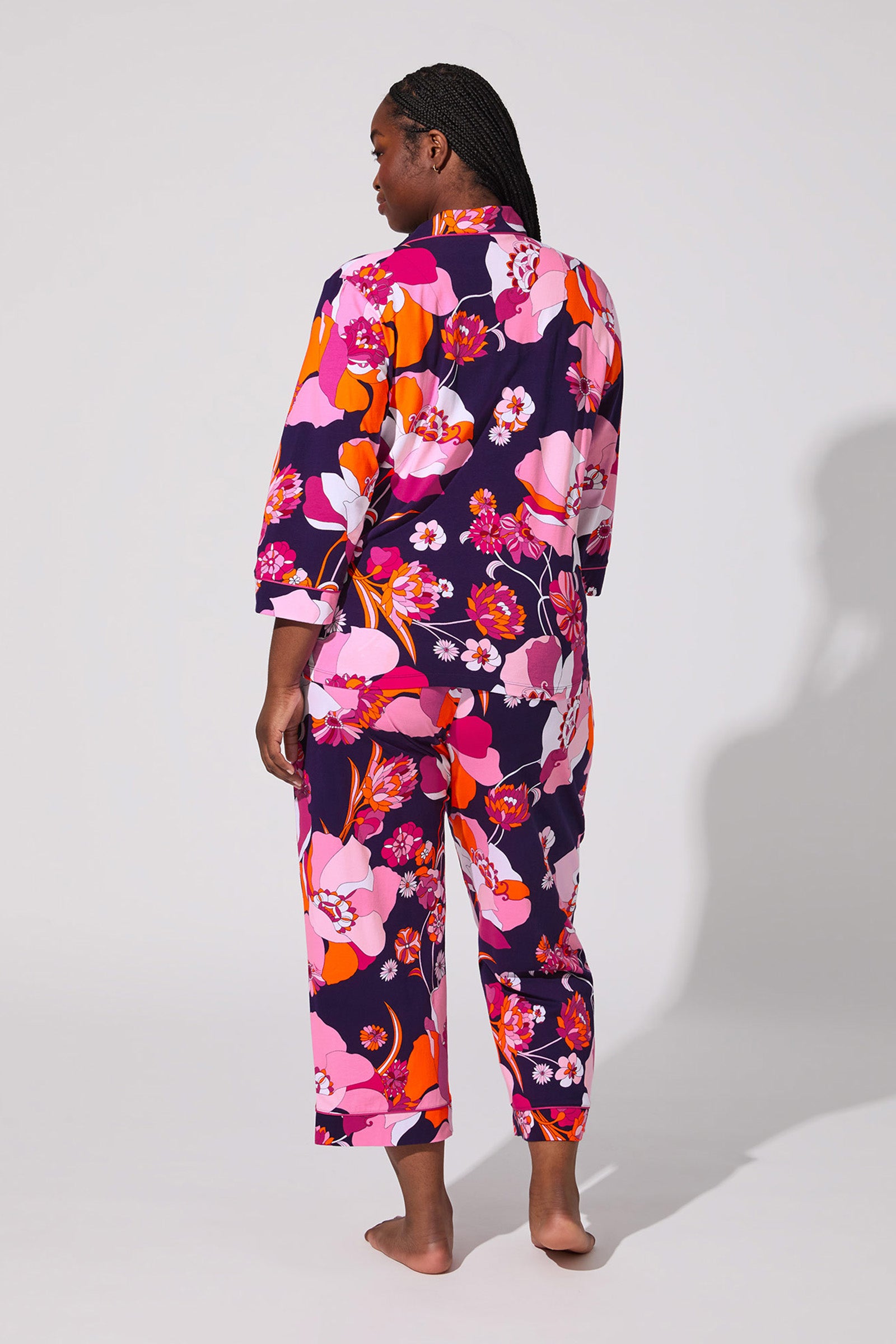 A person stands barefoot facing away, wearing the Floral Medley Crop Pj Set with a bold floral pattern in pink, orange, red, and purple. The background is plain light gray.