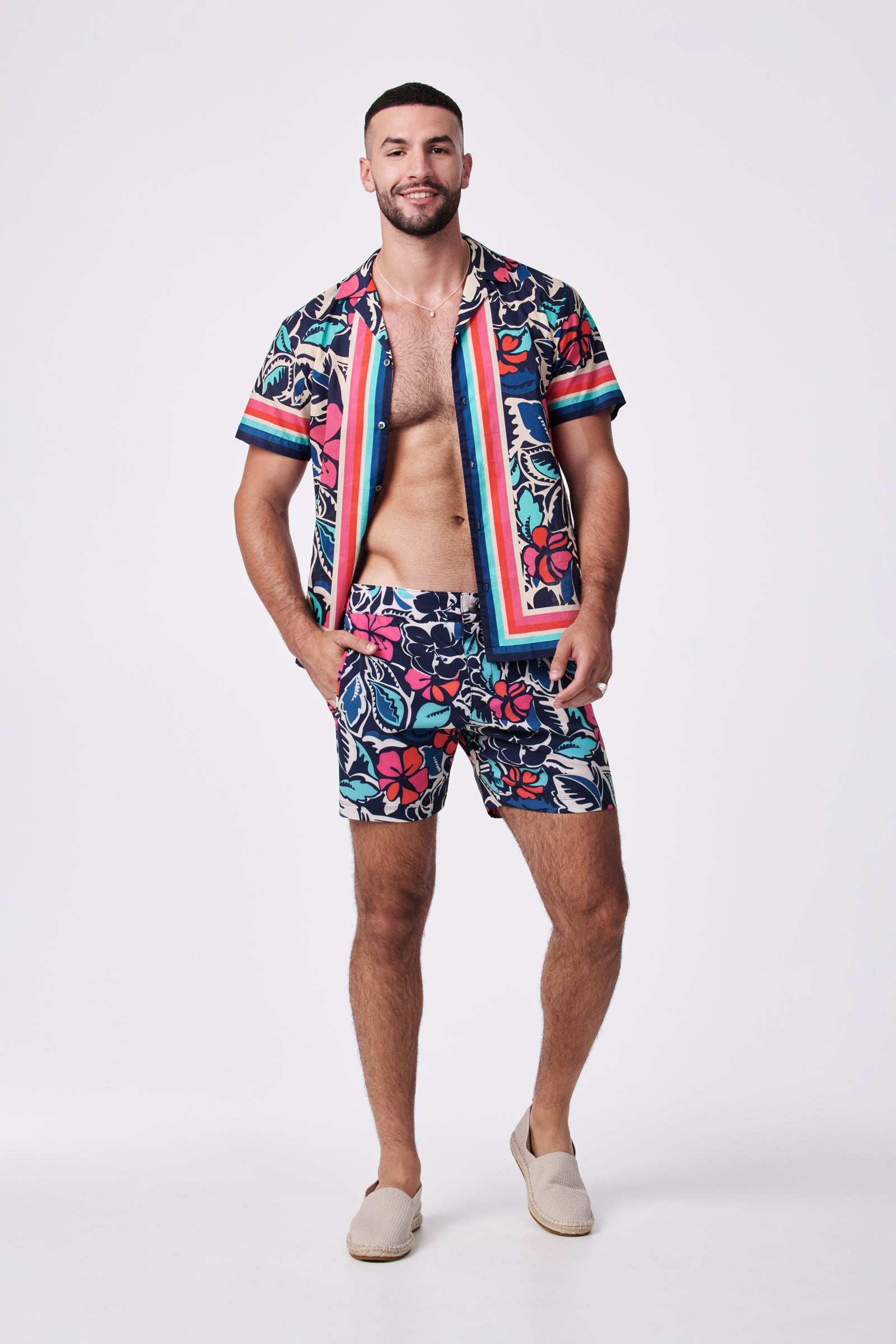 A man with facial hair smiles barefoot, wearing the Honolulu Board Short - Copacabana, a colorful tropical short with floral patterns, posing against a plain white background.