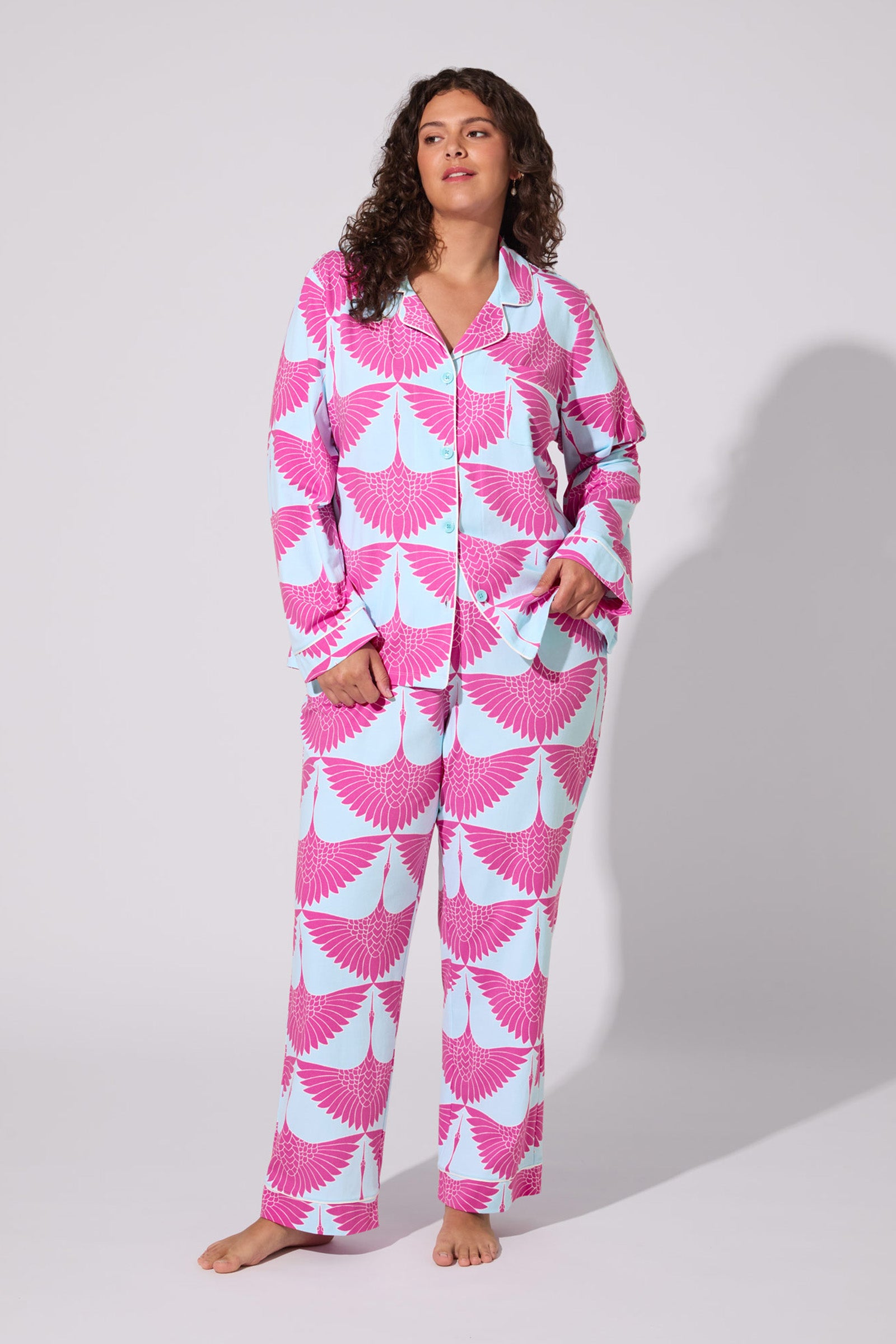A woman with curly hair models the Cranes Classic Pj Set, featuring a pink and light blue geometric wing print. She stands barefoot against a plain background, looking to the side with one hand in her pocket.