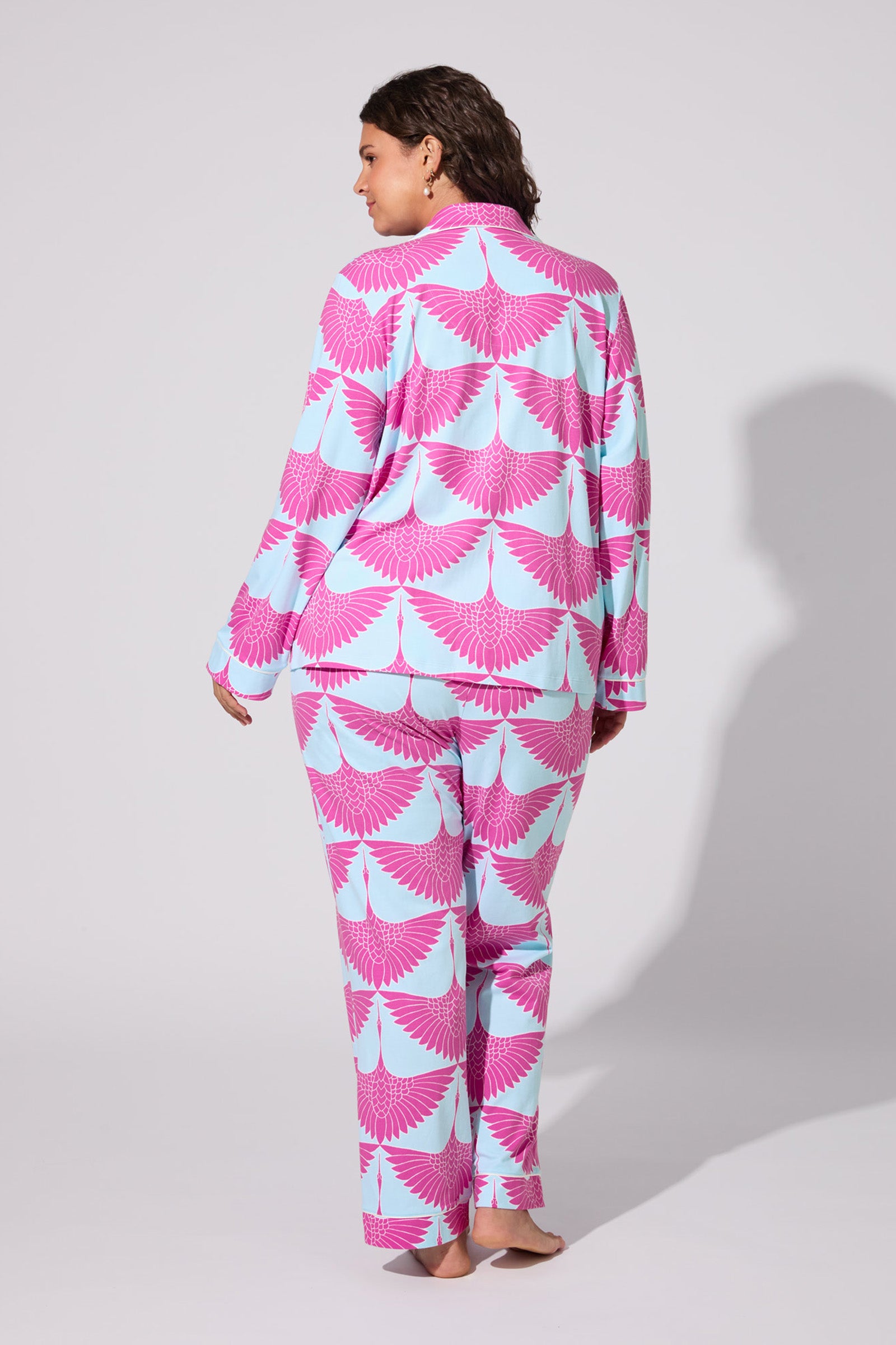 A woman stands barefoot, facing away from the camera, wearing the Cranes Classic Pj Set with a pink and light blue bird wing pattern. The background is plain light gray.