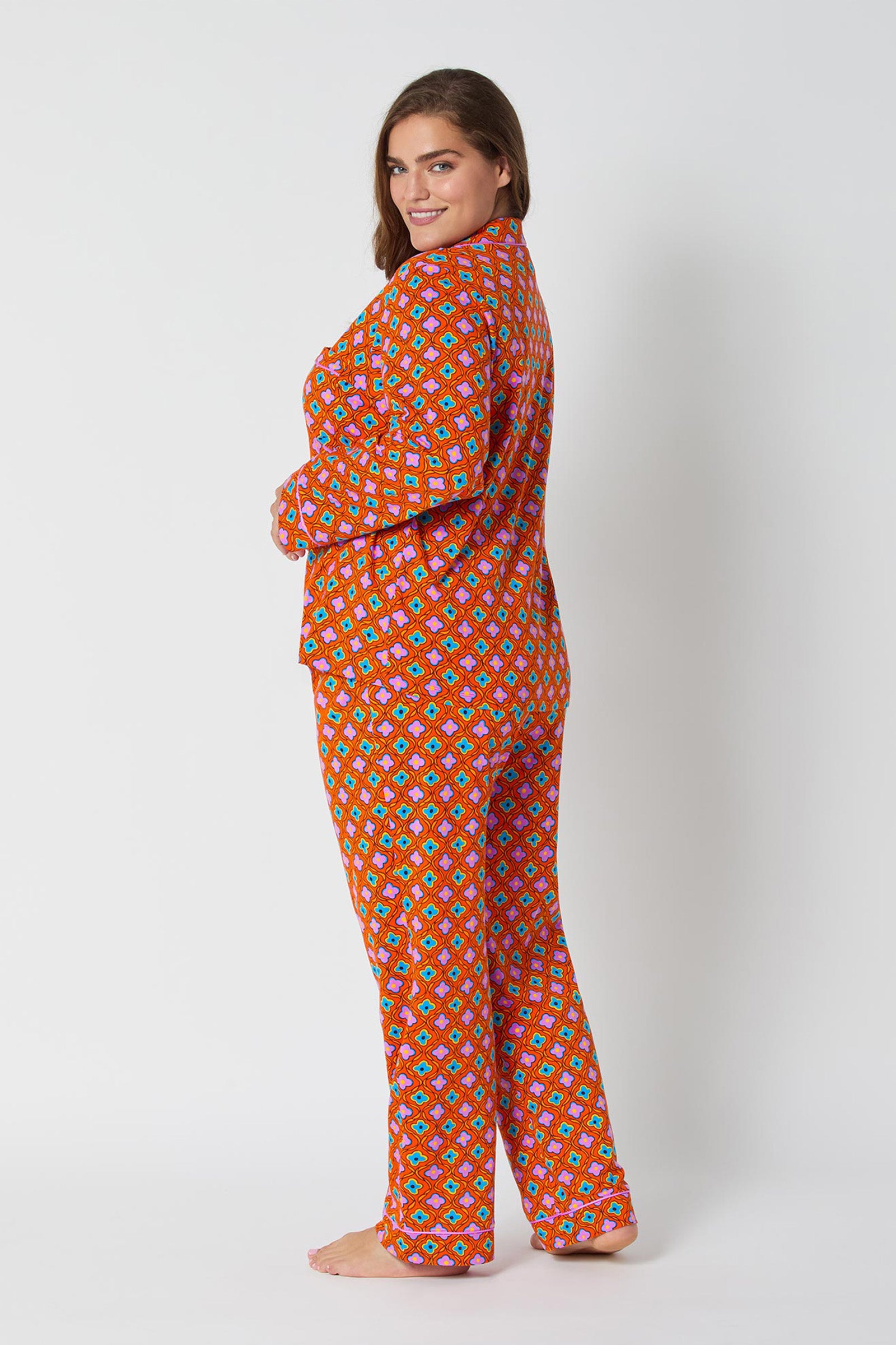 A woman stands barefoot against a white background, smiling over her shoulder. She is wearing the Daisy Foulard L/S Classic Pj, featuring colorful, loose-fitting pajamas with a vibrant geometric pattern.