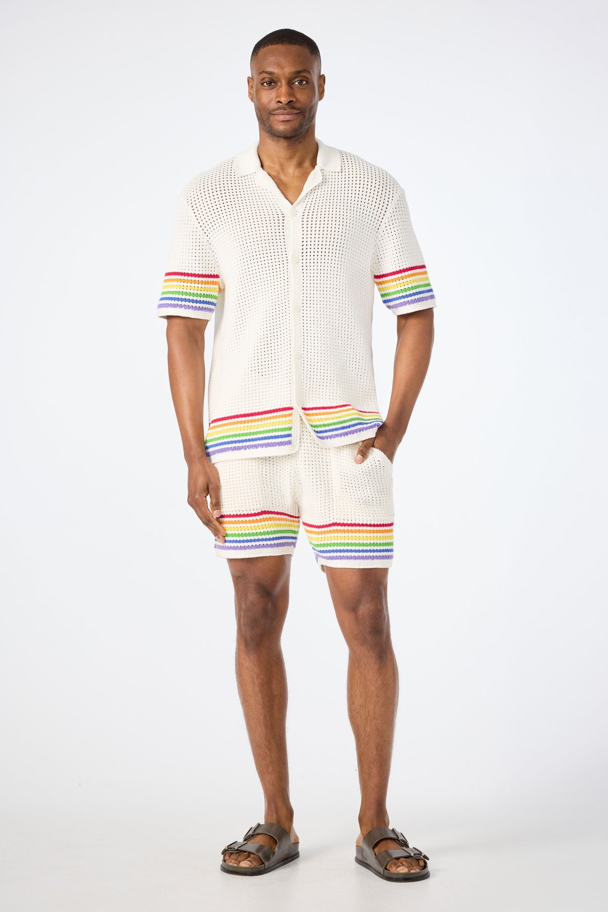 A man smiles in the Easton Short-Sleeve Resort Shirt - Pride, a cream knit top with colorful rainbow stripes on the sleeves and hem, paired with matching shorts. He wears brown sandals and poses against a plain white background.