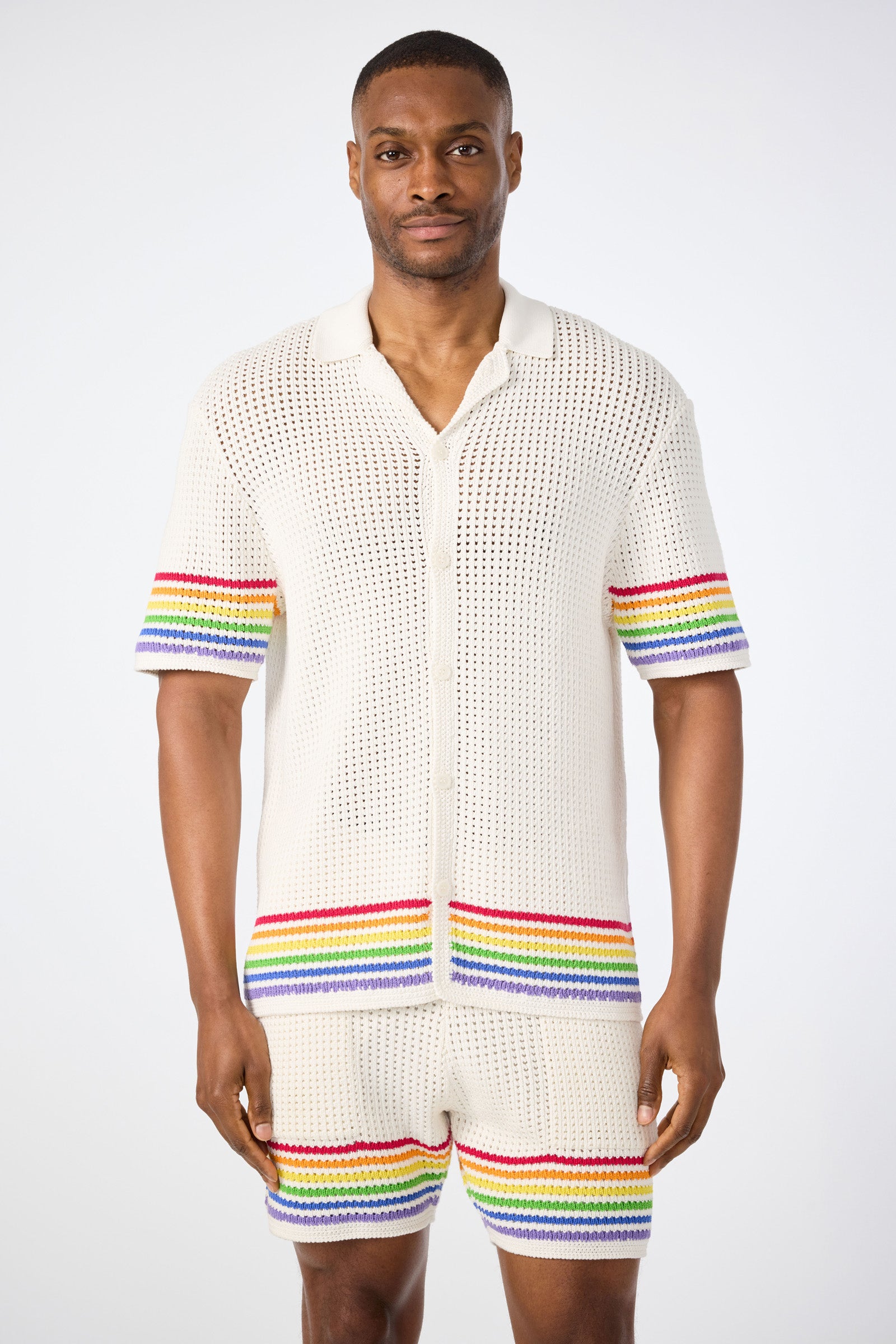 A man faces forward wearing the Easton Short-Sleeve Resort Shirt - Pride in multi, featuring horizontal rainbow stripes near the sleeves and hem. He pairs it with matching shorts; both are set against a plain white background.