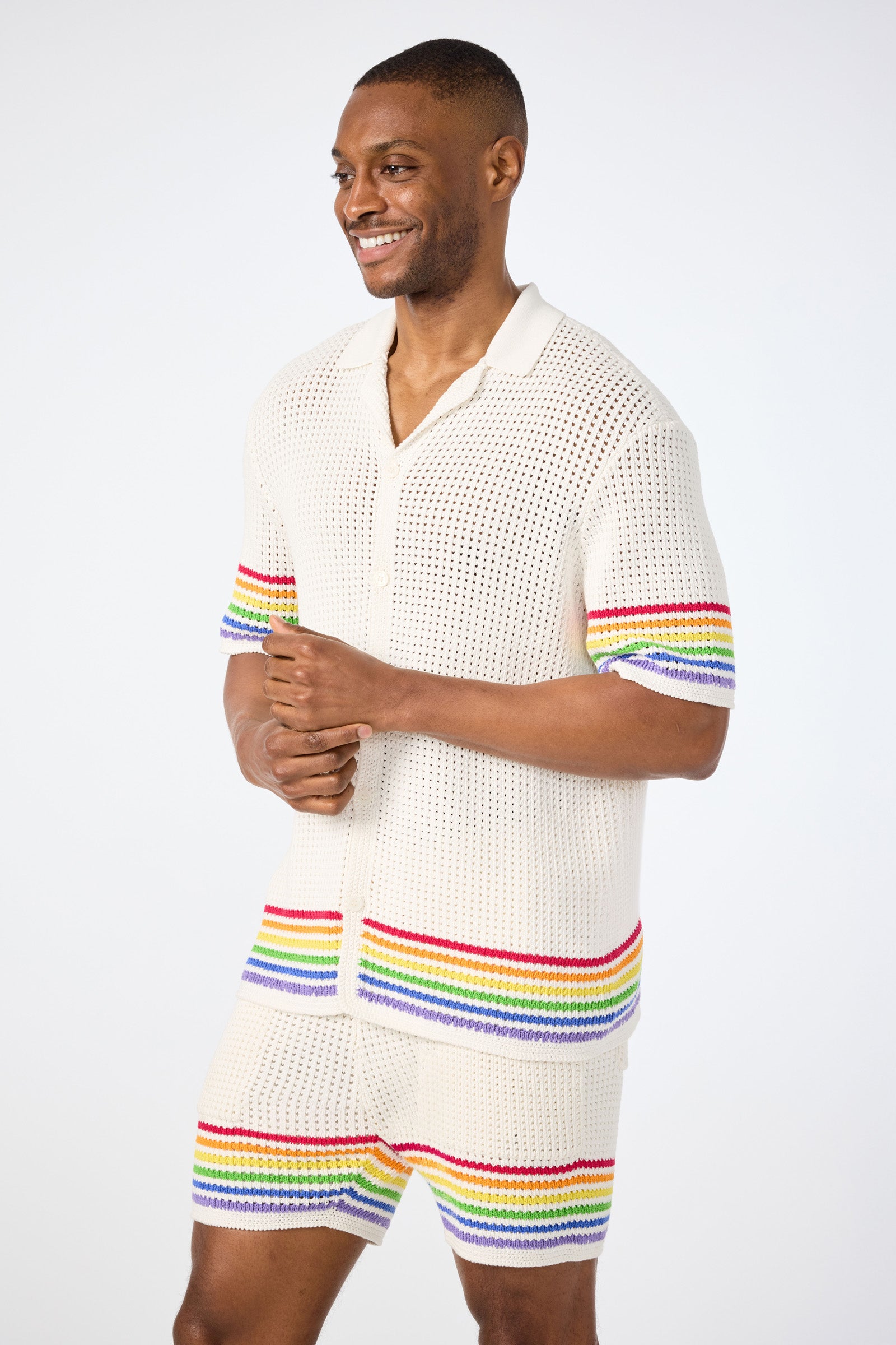 A smiling man wears the Easton Short-Sleeve Resort Shirt - Pride, a cream crochet shirt with colorful rainbow stripes on the sleeves and hem, paired with matching shorts, against a plain light background.