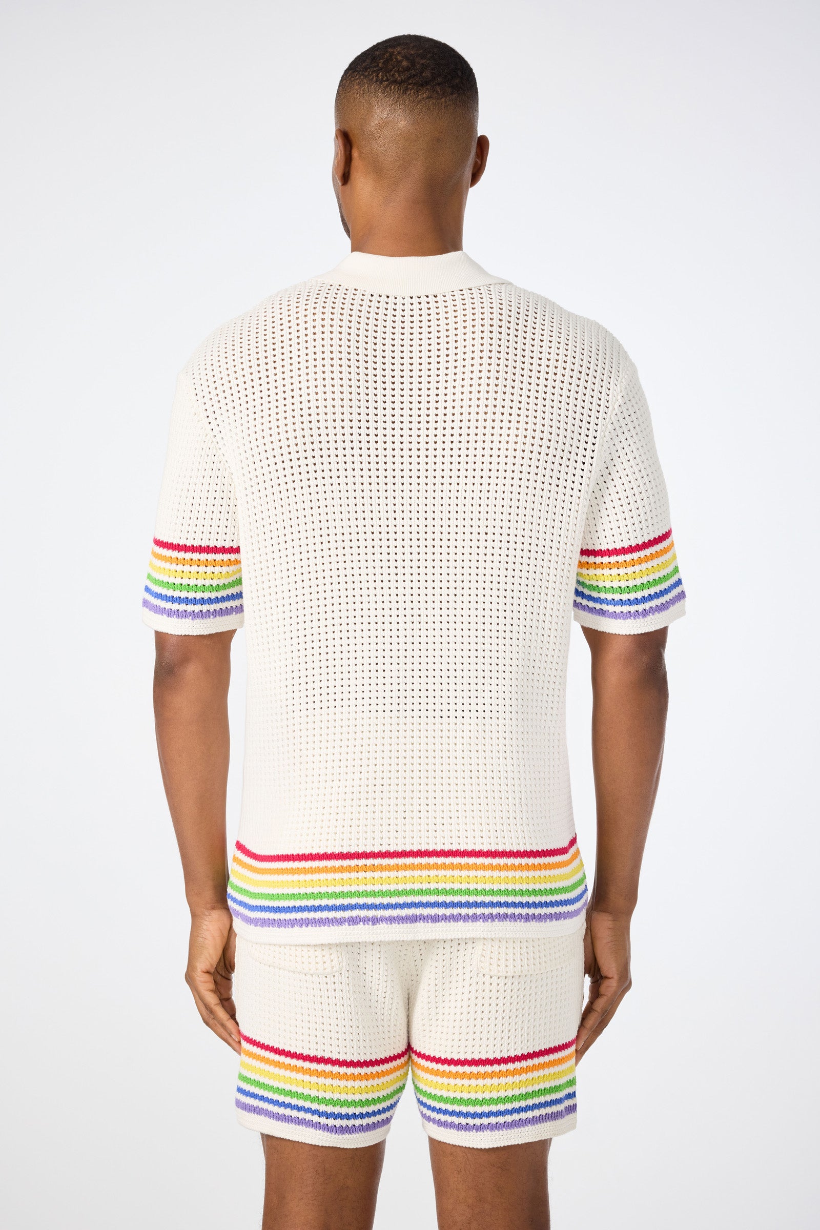 A person stands facing away, wearing the Easton Short-Sleeve Resort Shirt - Pride, a white crochet top with colorful rainbow stripes on the sleeves and hem. The background is plain white.