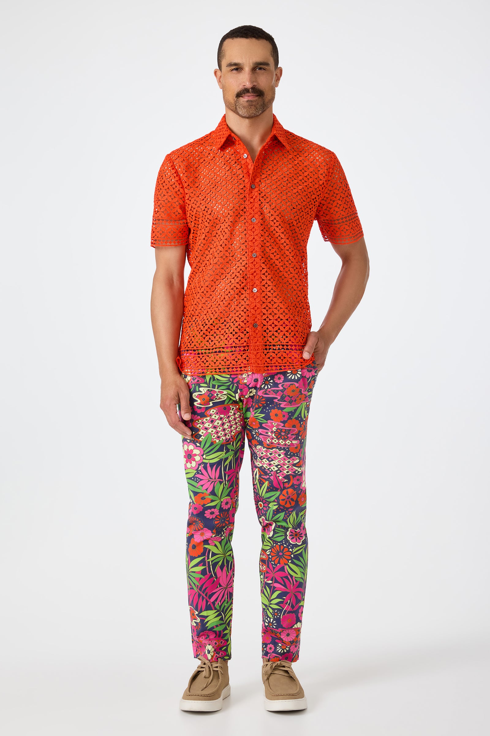 A man with short hair and a mustache stands against a white background wearing the bright orange Belmont Shore Shirt with a cutout pattern, colorful floral pants, and tan shoes.