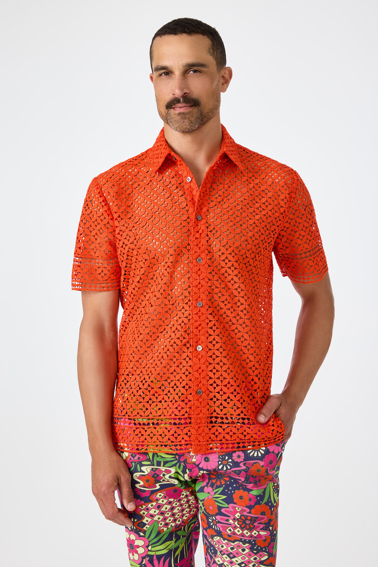 A man with short dark hair and a mustache wears the Belmont Shore Shirt in Ryuji Red—a bright, short-sleeved, see-through eyelet shirt—paired with colorful, floral pants, standing against a plain white background.
