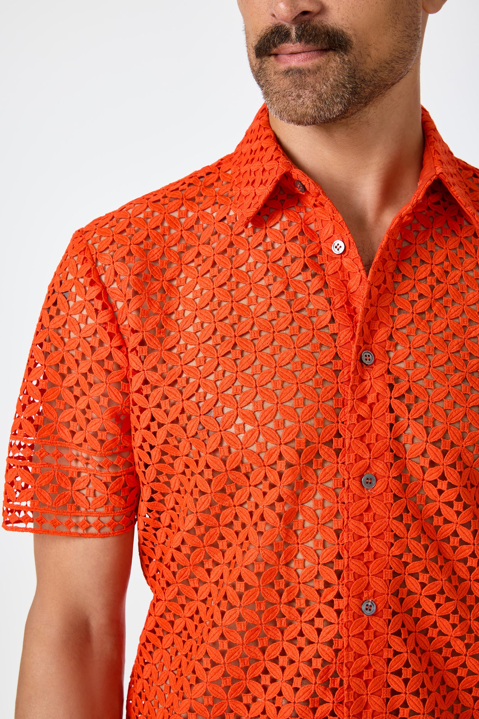 A man with a mustache wears the Belmont Shore Shirt—a bright orange, short-sleeved, button-up featuring a geometric see-through lace pattern. The image is cropped at his upper chest and mouth.