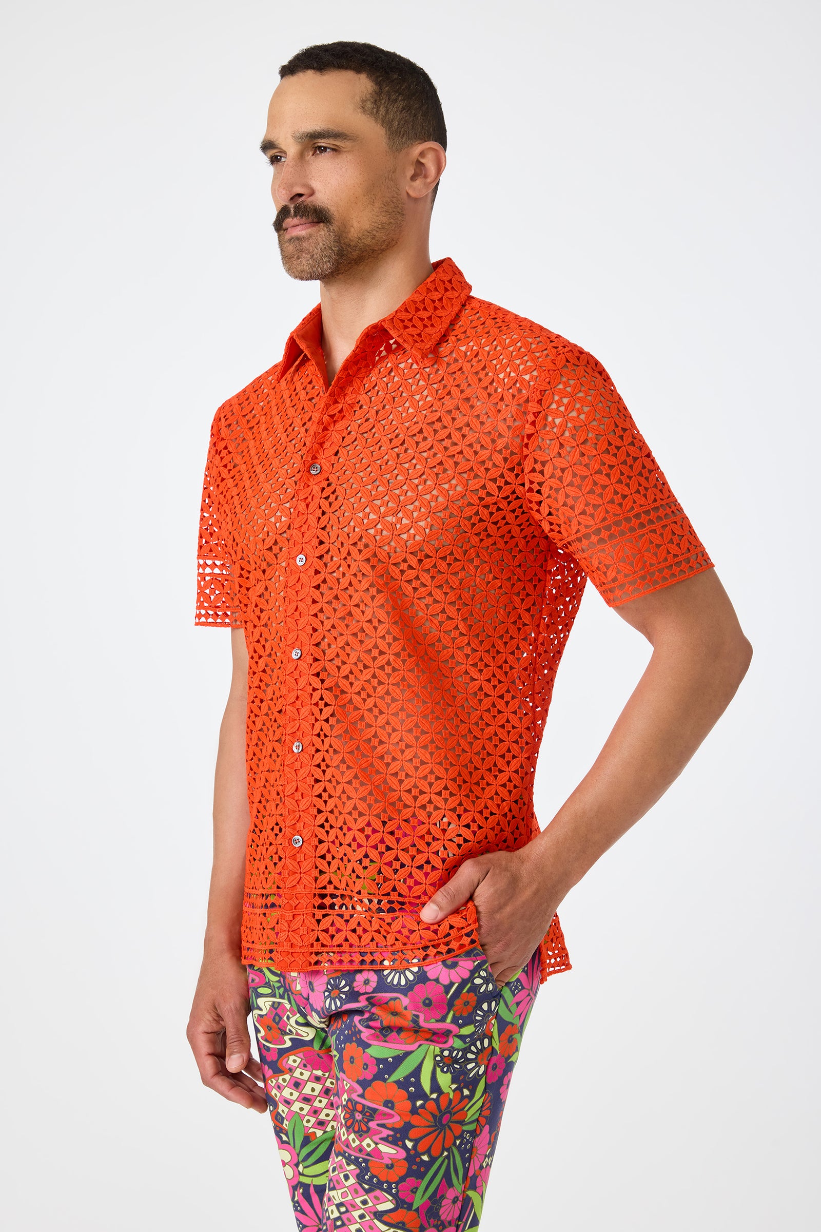 A man with a short beard poses against a white background, wearing the Belmont Shore Shirt—a bright orange, short-sleeved, see-through lace shirt—and colorful floral pants, with one hand in his pocket.