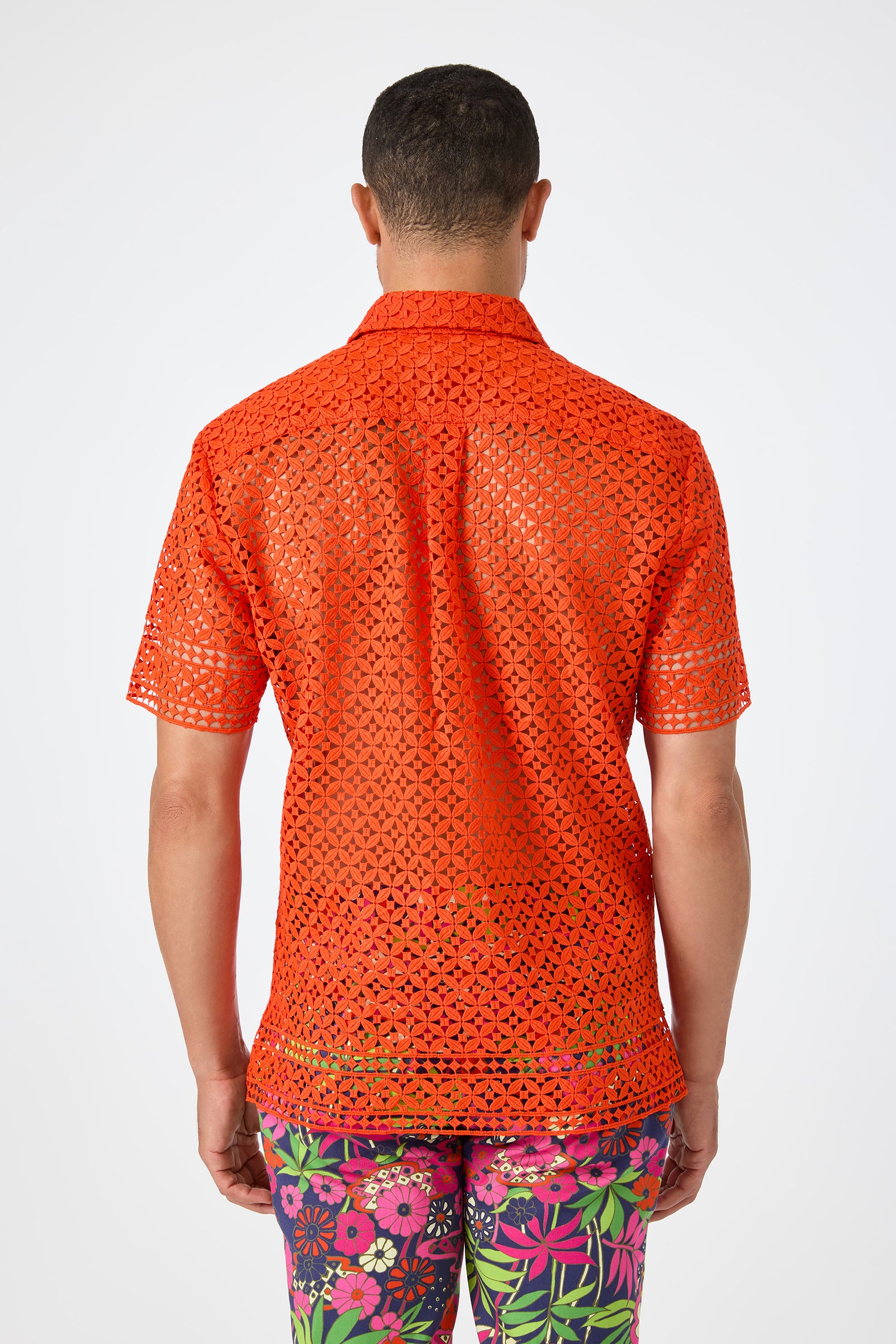 A person is shown from the back wearing the Belmont Shore Shirt, a bright orange, short-sleeve, see-through crochet top, paired with colorful floral pants against a plain light background.