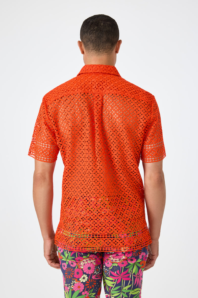 A person is shown from the back wearing the Belmont Shore Shirt, a bright orange, short-sleeve, see-through crochet top, paired with colorful floral pants against a plain light background.