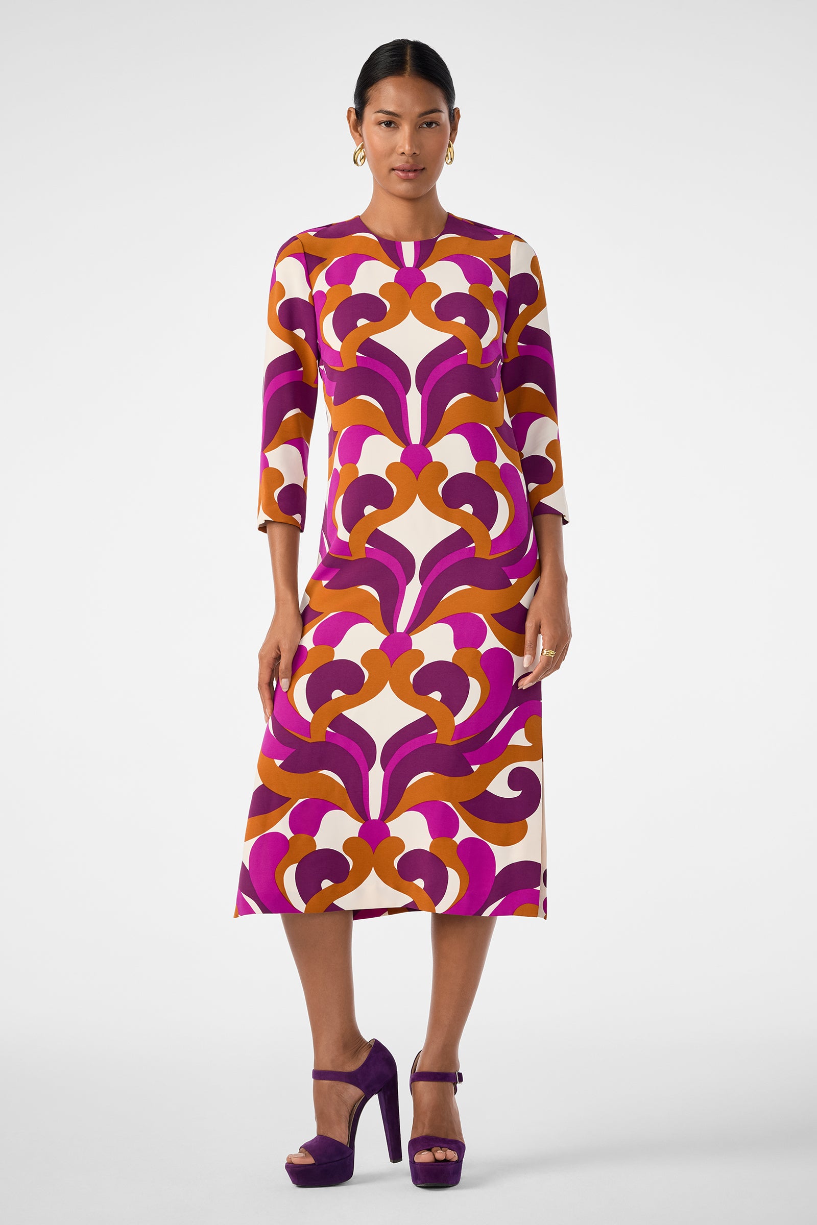 A woman models the Cointreau 2 Dress—a long-sleeved, mid-length dress with a bold geometric pattern in purple, orange, and cream—paired with purple platform heels and gold hoop earrings against a white background.