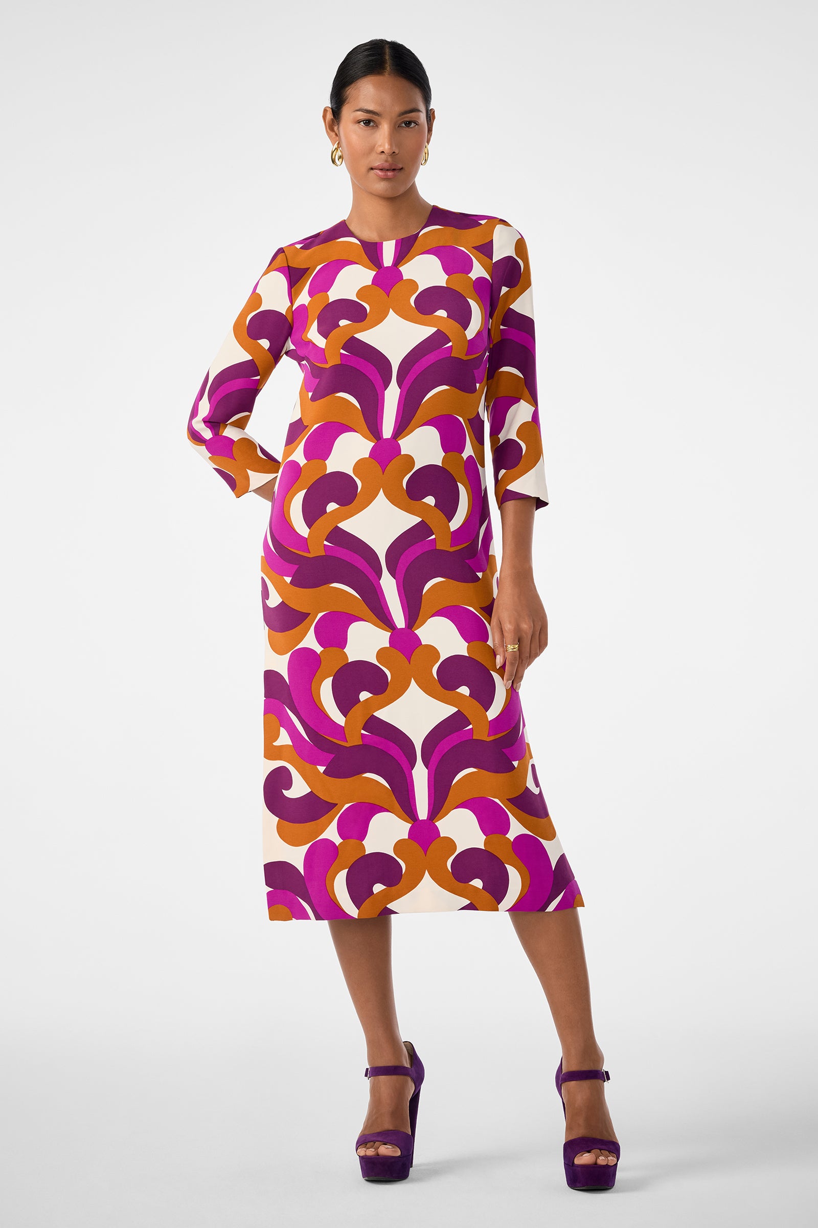 A woman stands confidently in the Cointreau 2 Dress, a long-sleeve, midi-length piece featuring a vibrant geometric pattern in pink, orange, and purple, paired with purple platform heels.