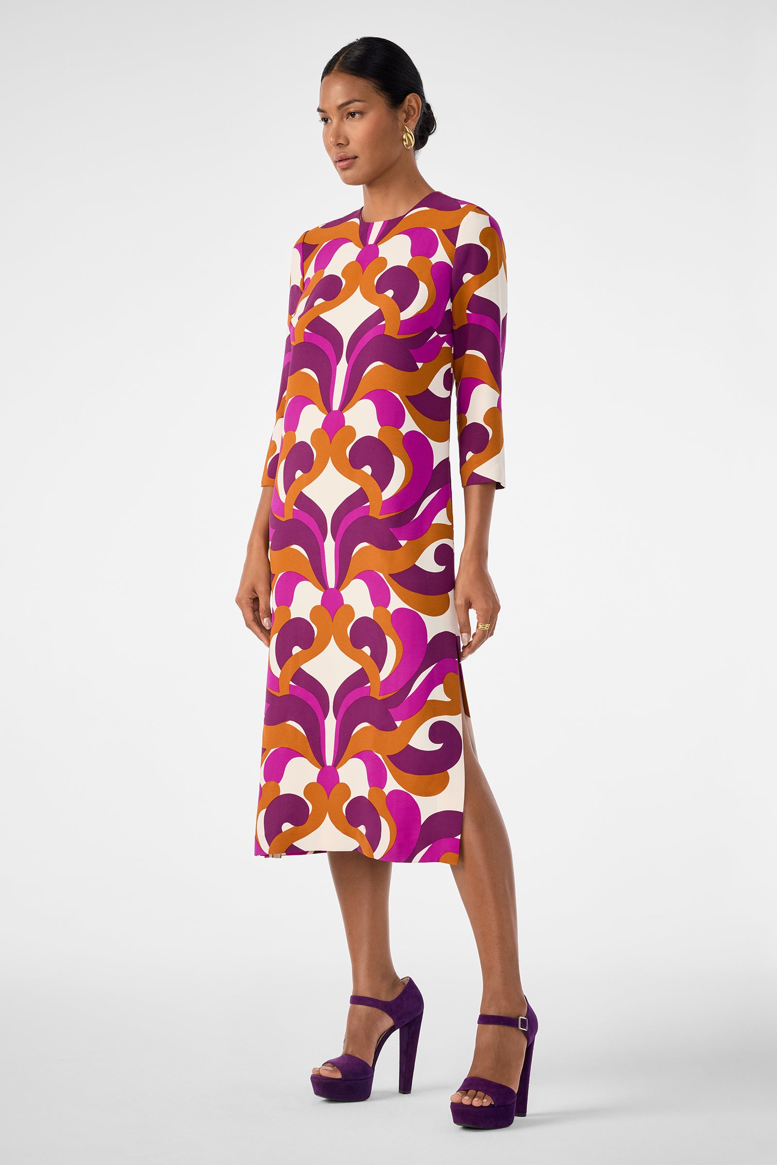 A woman models the Cointreau 2 Dress, a colorful midi with orange, pink, and purple swirls, three-quarter sleeves, and side slit; styled with purple platform heels against a plain background.