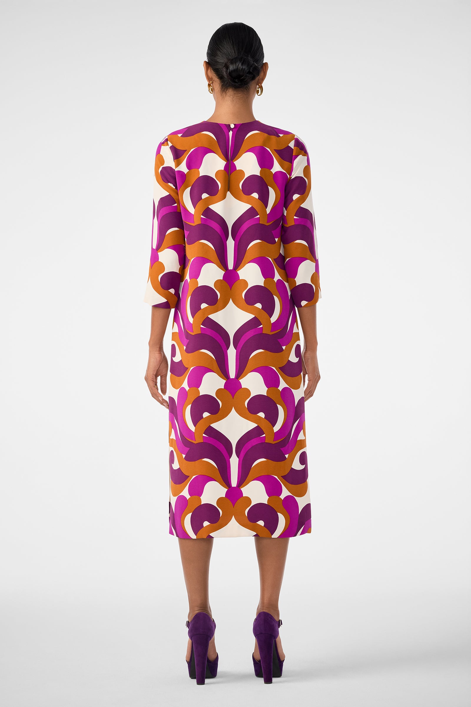 A woman faces away, wearing the Cointreau 2 Dress—a knee-length piece with a bold retro purple, orange, and white pattern. Her hair is in a neat bun, and she sports matching purple high heels. The background is plain white.
