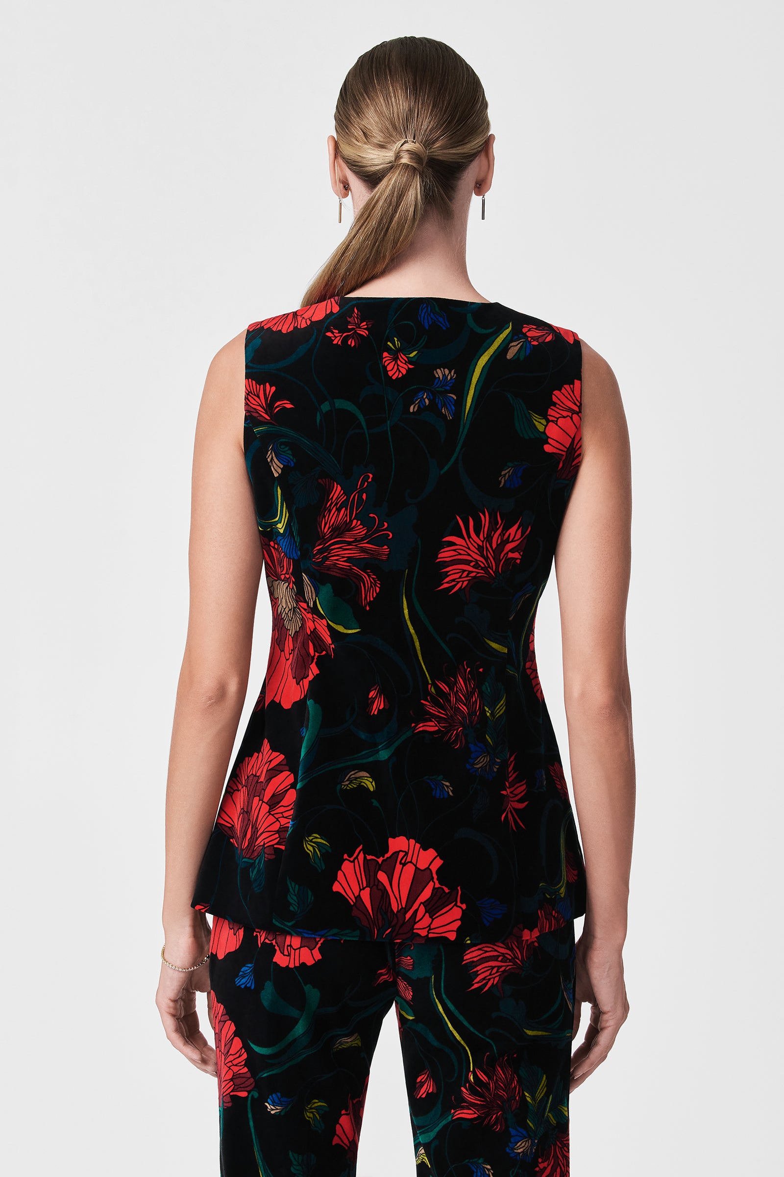A woman with light skin and straight, light brown hair in a low ponytail is seen from the back wearing the Kary Printed Velvet Vest, a sleeveless black top with bold red and green floral patterns.