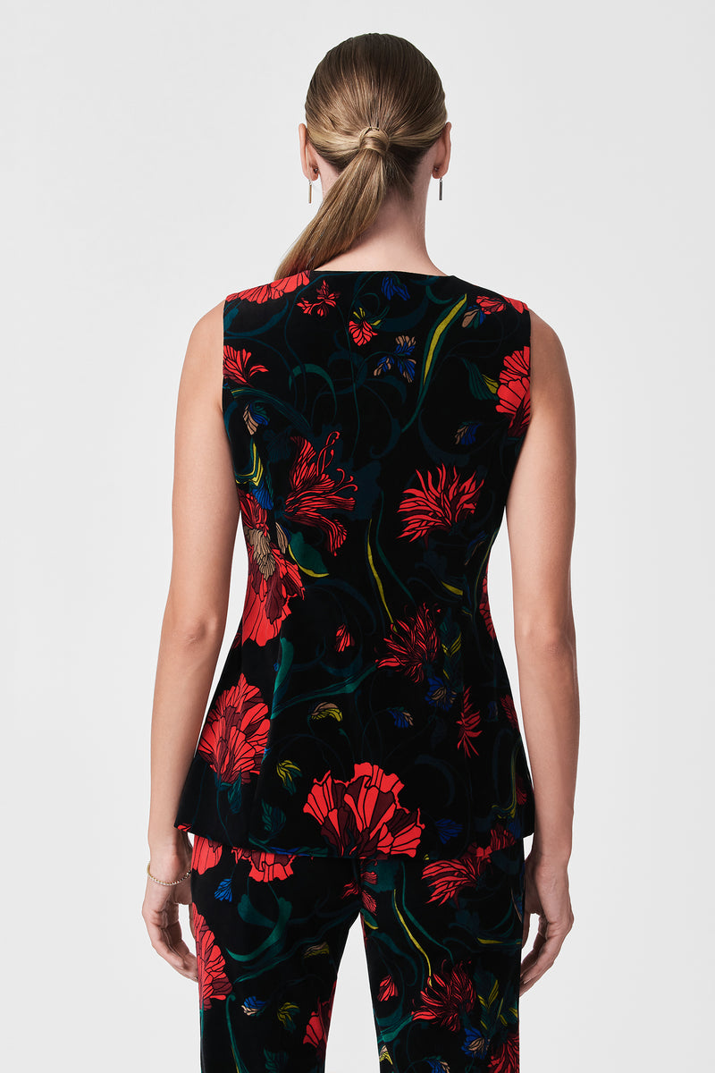 A woman with light skin and straight, light brown hair in a low ponytail is seen from the back wearing the Kary Printed Velvet Vest, a sleeveless black top with bold red and green floral patterns.