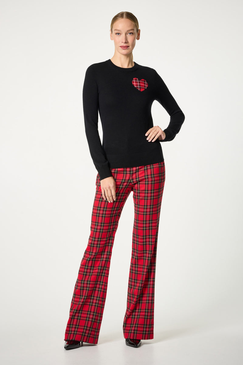 A woman with her hair tied back stands against a white background wearing the Plaid Heart Crewneck Sweater, which features a red plaid heart on the chest, paired with red plaid flared pants and black pointed shoes.