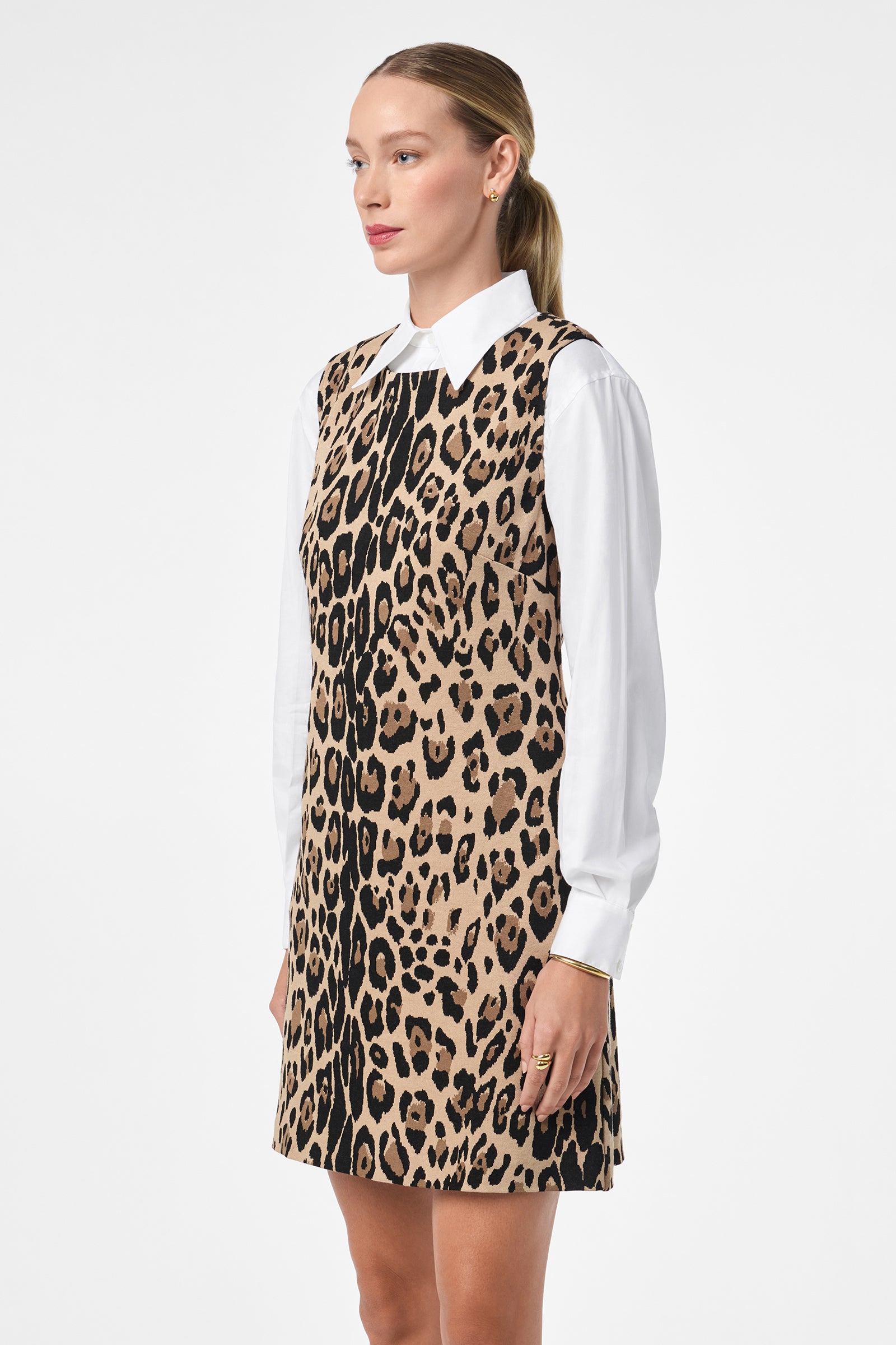 A woman with straight, light brown hair pulled back models the Adapted Dress—a sleeveless tan and black leopard print style—layered over a white collared shirt against a plain white background.