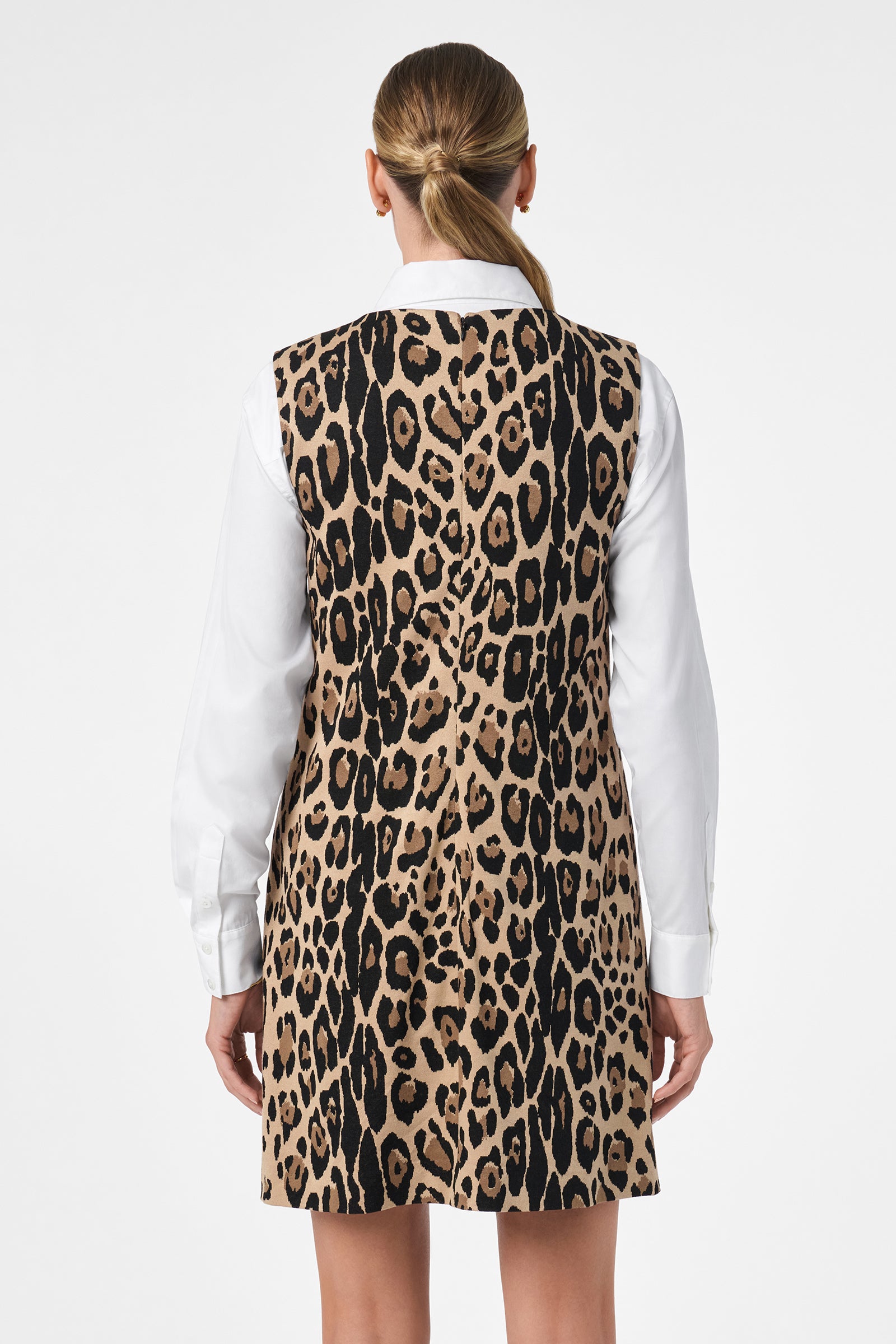 A woman with straight, blonde hair in a low ponytail is shown from behind wearing the Adapted Dress, a sleeveless, knee-length leopard print style layered over a white long-sleeve blouse. The background is plain white.