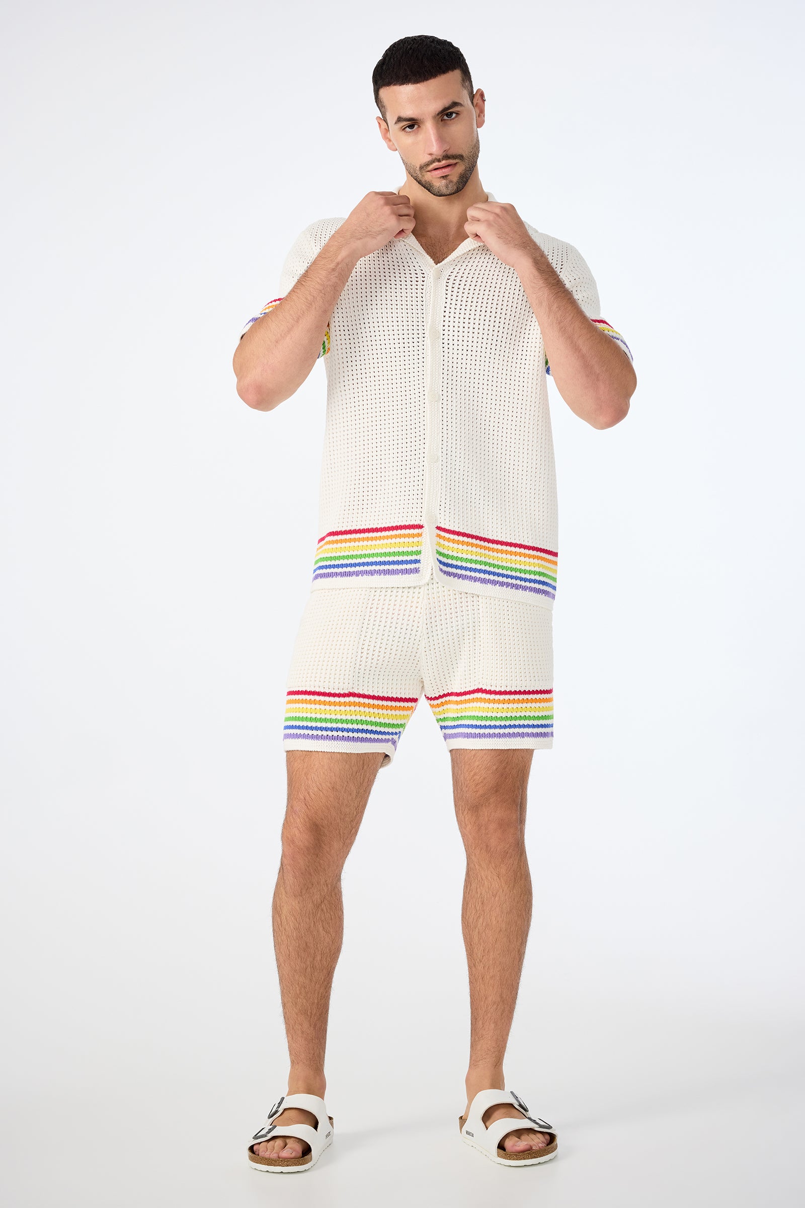 A man poses against a white background wearing the August Open-Knit Short - Pride, featuring white knit fabric with horizontal rainbow stripes, paired with a matching shirt and white sandals while adjusting his collar.