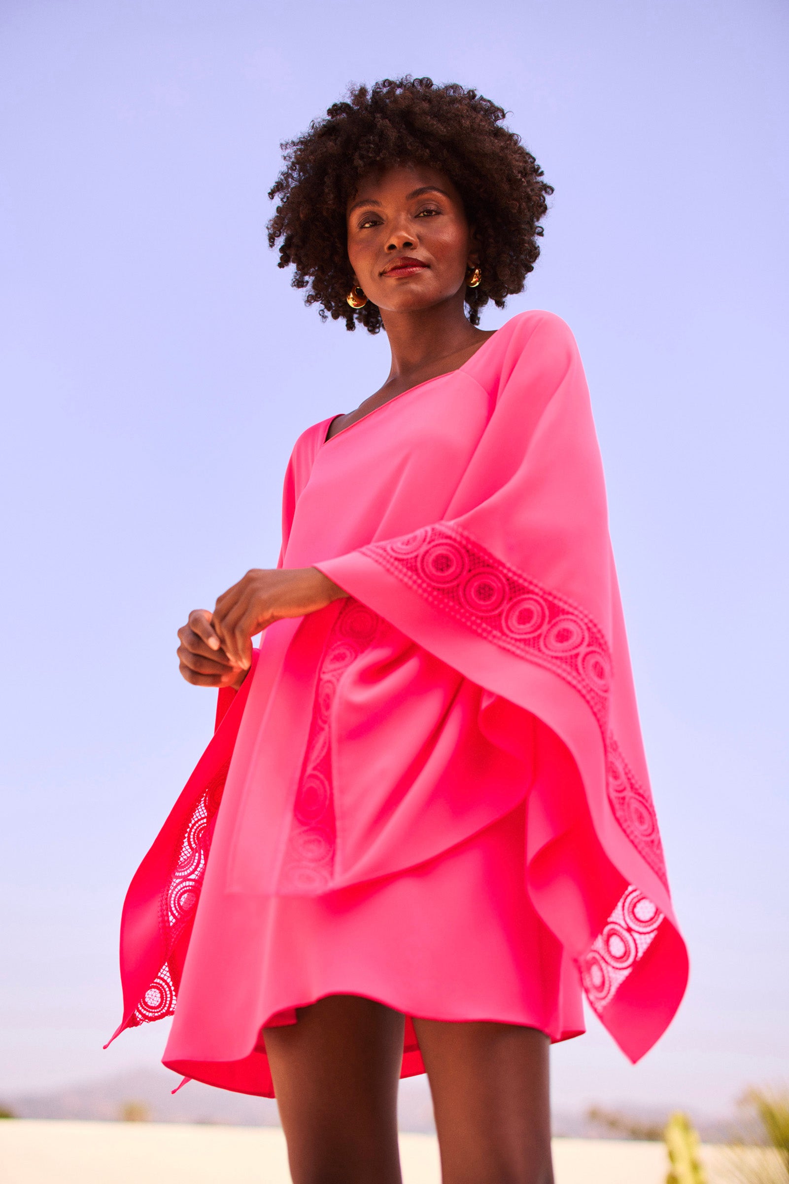 A woman with curly hair wears the Aqualane Side Flounce Dress, a vibrant pink, flowy outfit with patterned details, standing outdoors under a clear blue sky and looking confidently at the camera.