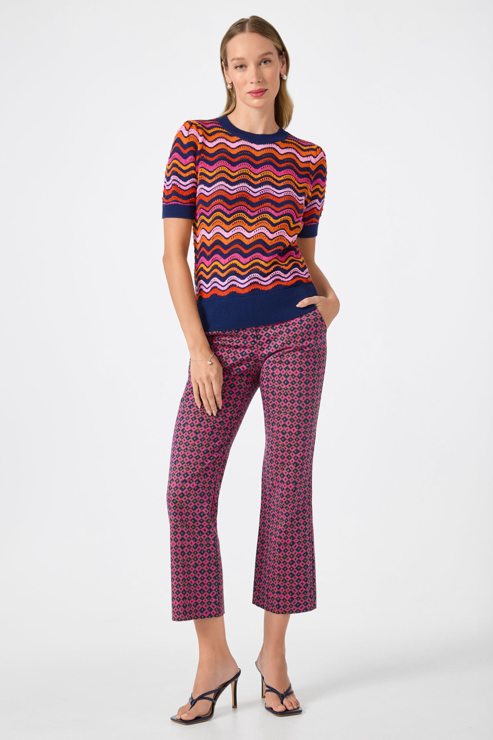 A woman stands against a white background wearing a colorful, short-sleeve, wavy-striped sweater with Lulu Cropped Pants in Himari Herringbone and black high-heeled sandals.