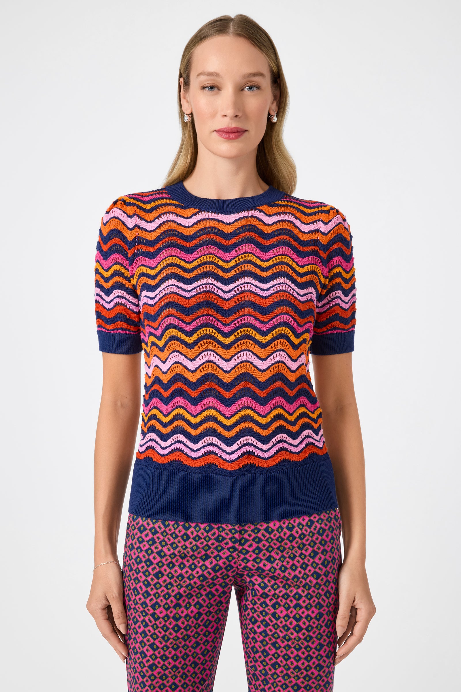 A woman faces forward wearing the Marbella Wavy Knit Pullover, a colorful short-sleeve sweater with wavy stripes in orange, pink, yellow, and blue, paired with patterned pink and purple pants. She has long straight hair and a neutral expression.