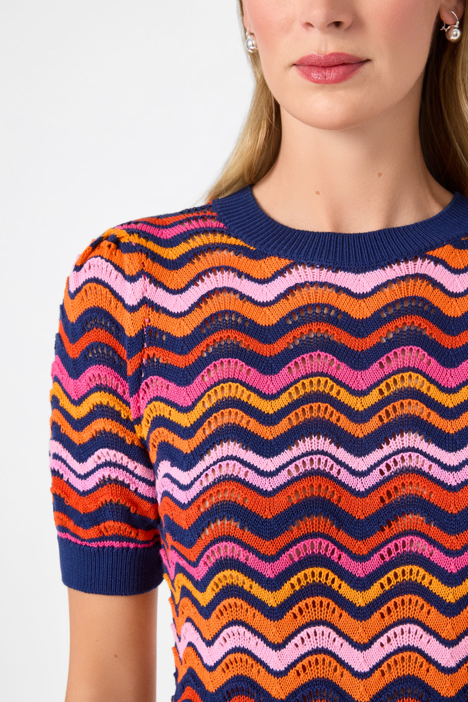 A woman models the Marbella Wavy Knit Pullover, a colorful short-sleeve top with pink, orange, red, and navy blue wavy stripes. The photo highlights the shirt and her light lipstick, showing her from the shoulders up.