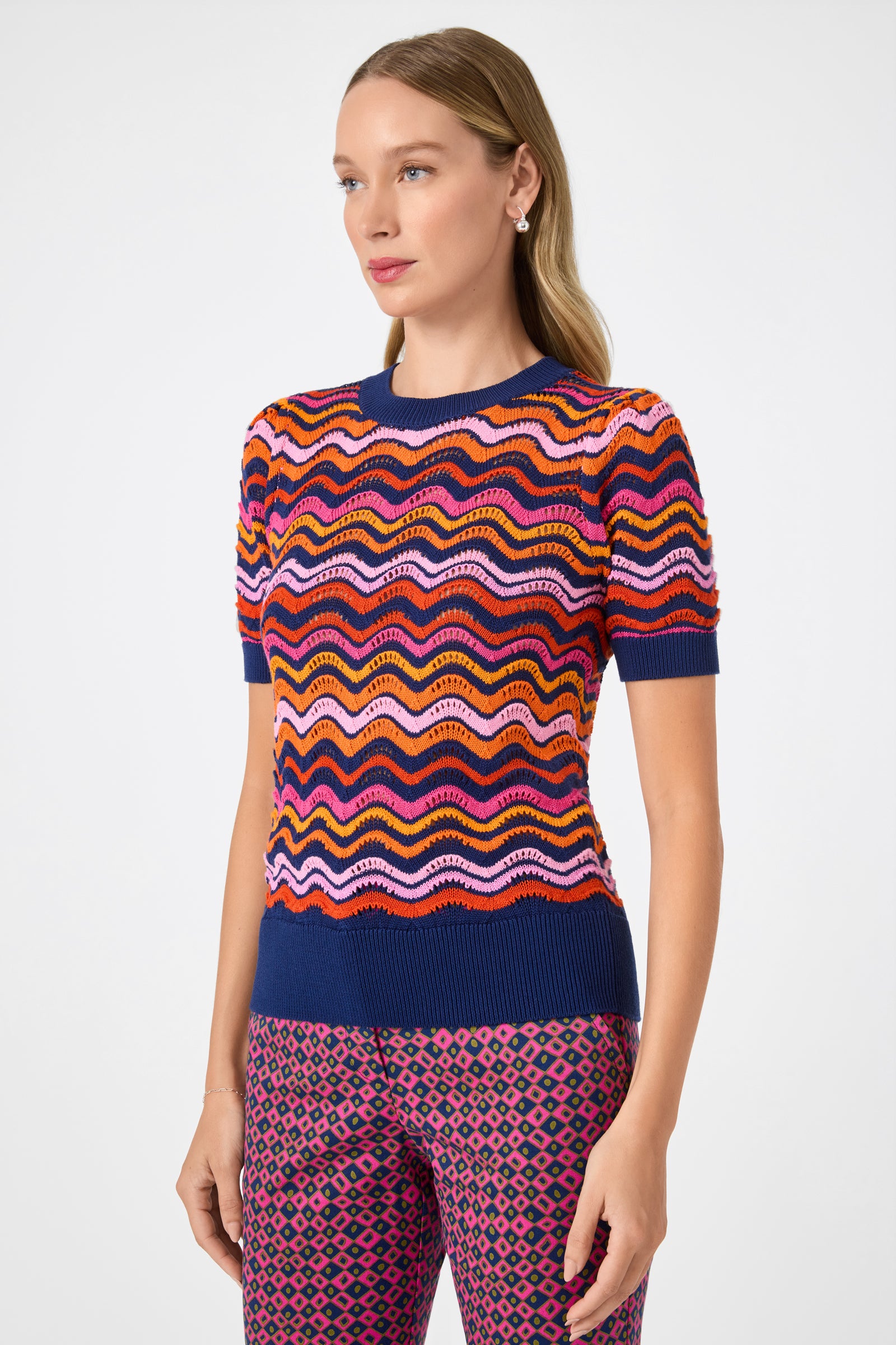A woman wears the Marbella Wavy Knit Pullover—a short-sleeve sweater with a multicolored zigzag pattern and blue trim—paired with pink and purple patterned pants, standing against a plain light background.
