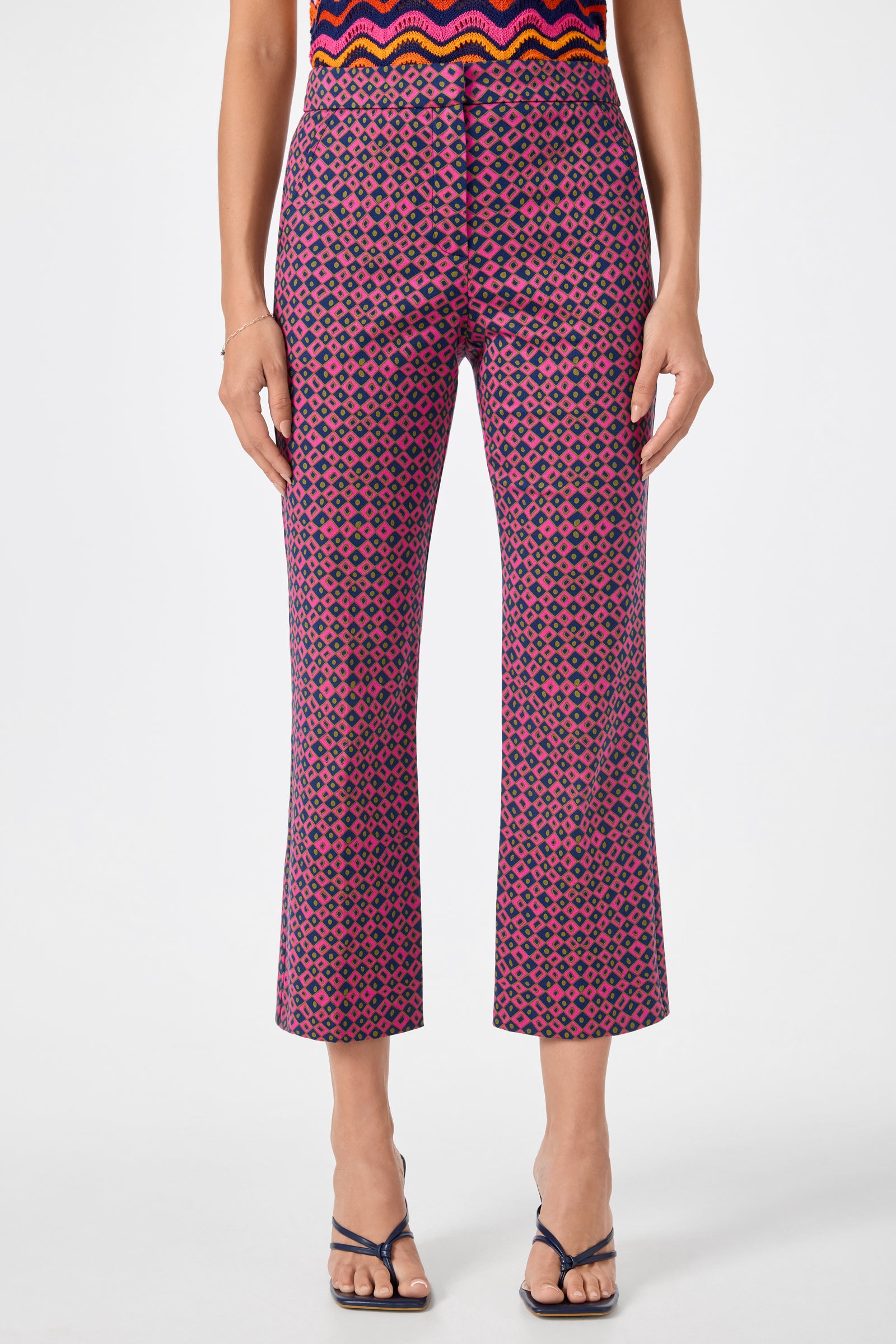 A person wearing the Lulu Cropped Pants - Himari Herringbone, featuring a high-waisted, geometric pink and navy pattern, paired with black strappy heeled sandals and a colorful top.
