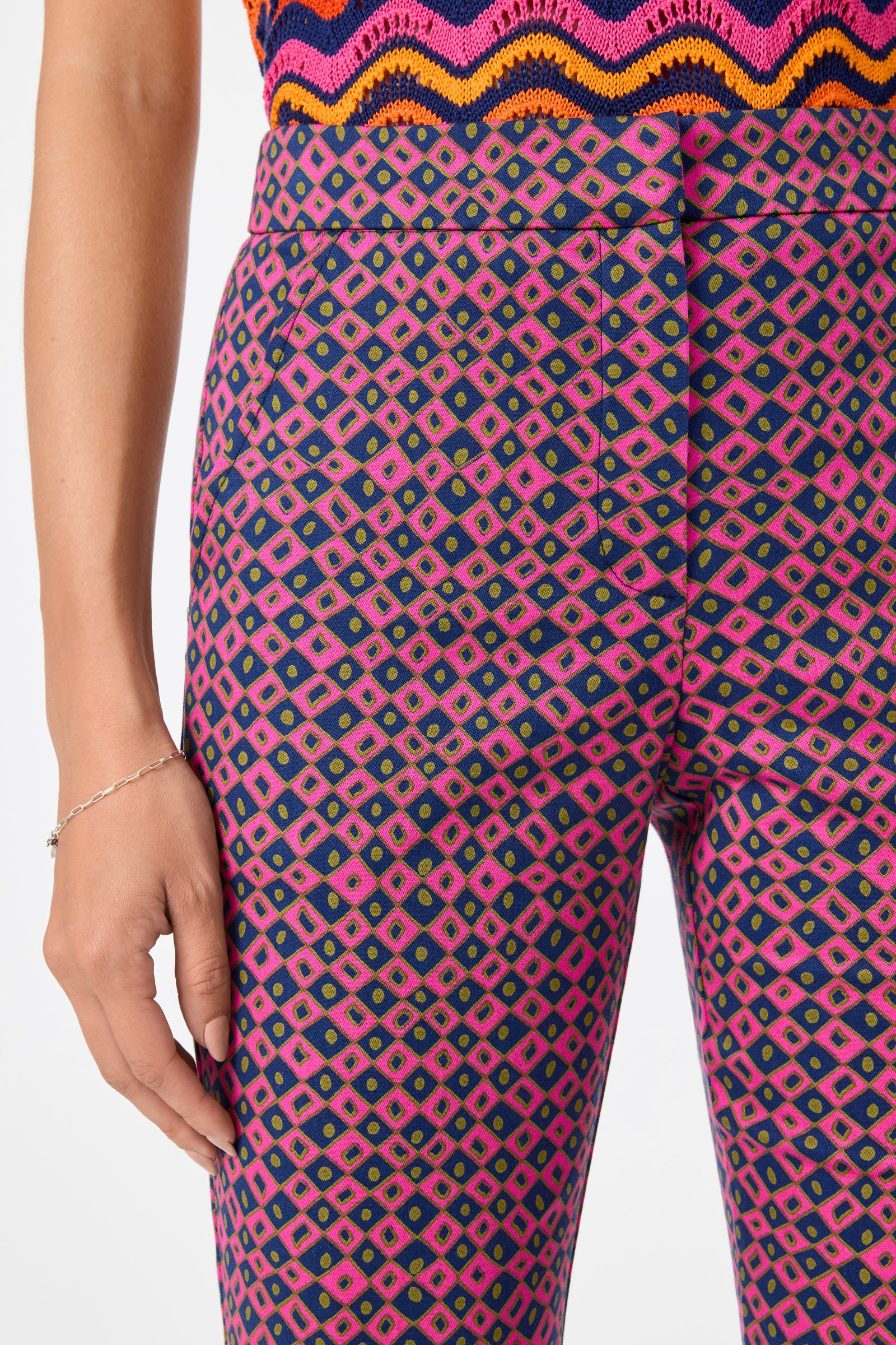 A person wears the Lulu Cropped Pants - Himari Herringbone, featuring a vibrant pink, purple, and green geometric pattern, paired with a colorful wavy-striped knit top; their left hand rests by their side.