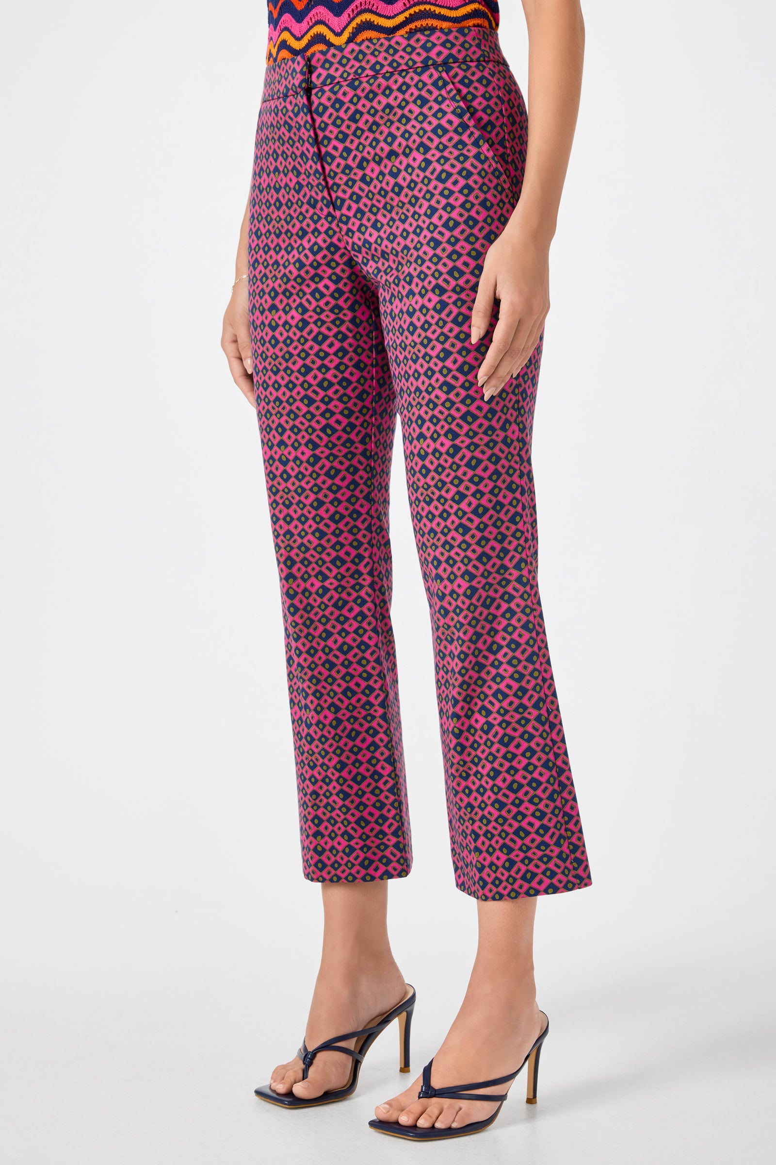 A person wears Lulu Cropped Pants - Himari Herringbone, featuring a pink and blue geometric pattern, paired with a colorful patterned top and black high-heeled sandals. The background is plain white.