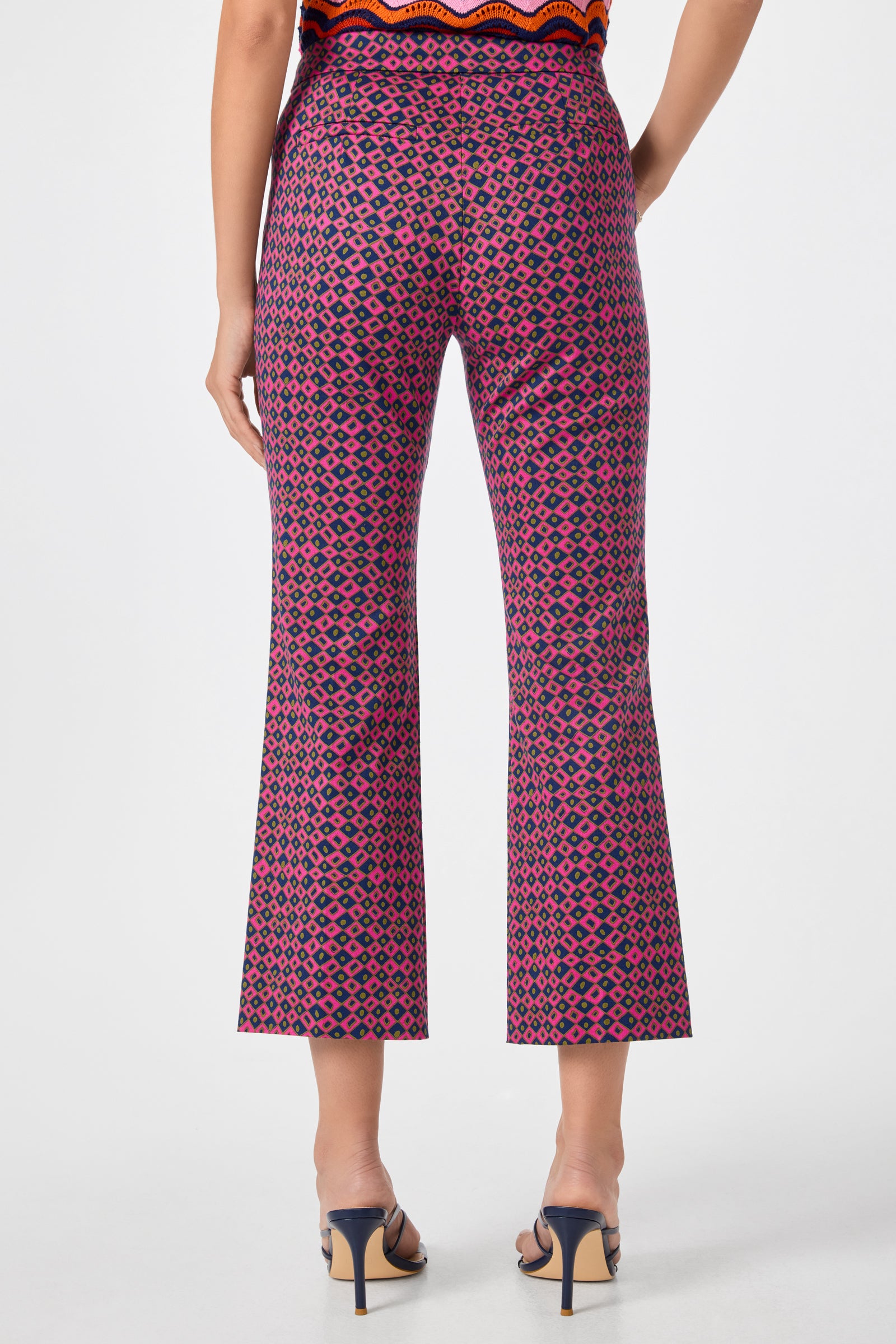 A woman faces away from the camera, wearing Lulu Cropped Pants - Himari Herringbone with a pink and black geometric pattern, black heeled sandals, and a partially visible orange patterned top.