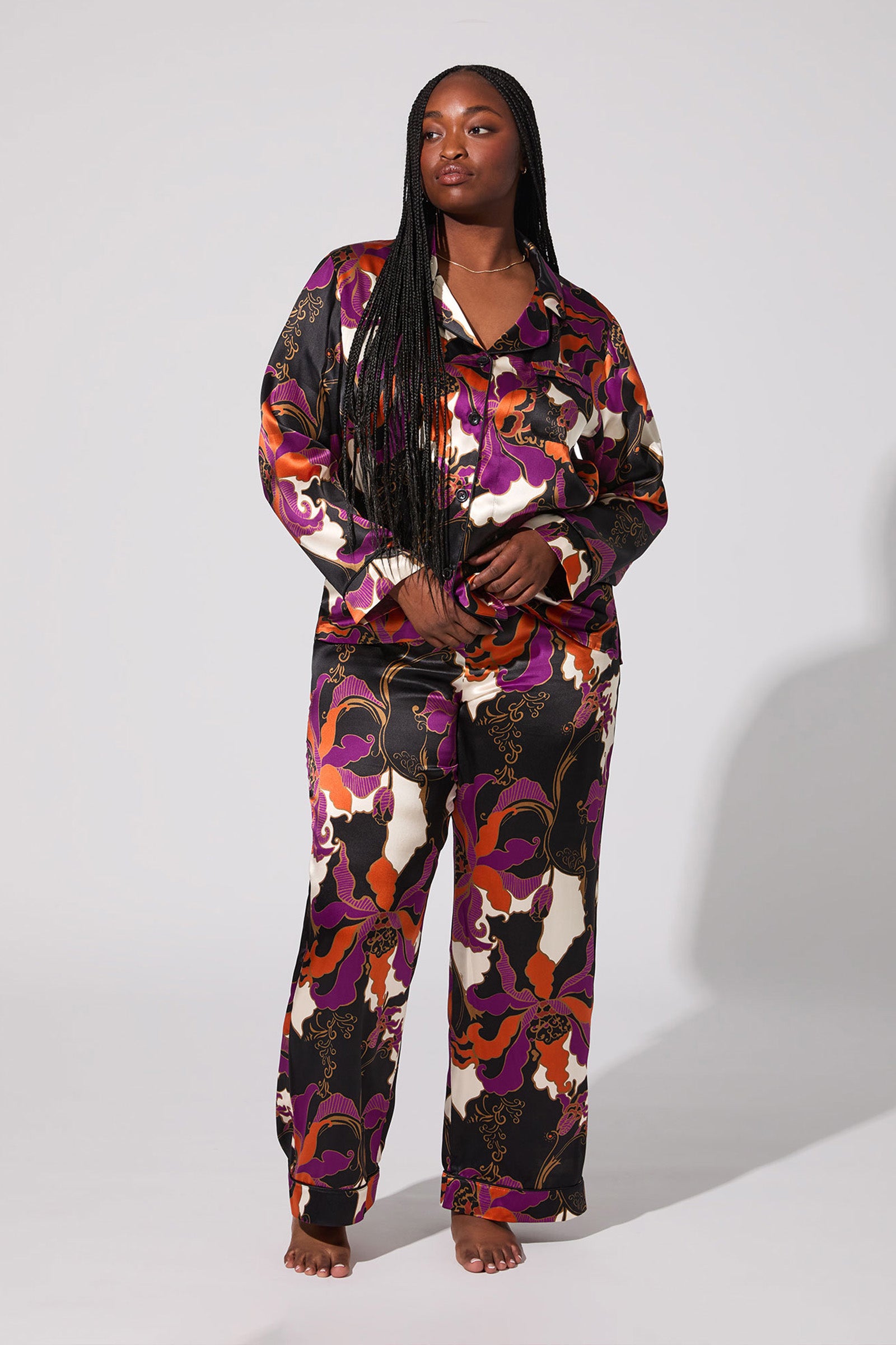 A woman with long braided hair stands barefoot and confidently in the Fall Floral Classic Silk Pj, featuring a bold purple, orange, red, black, and cream floral pattern, against a plain background.
